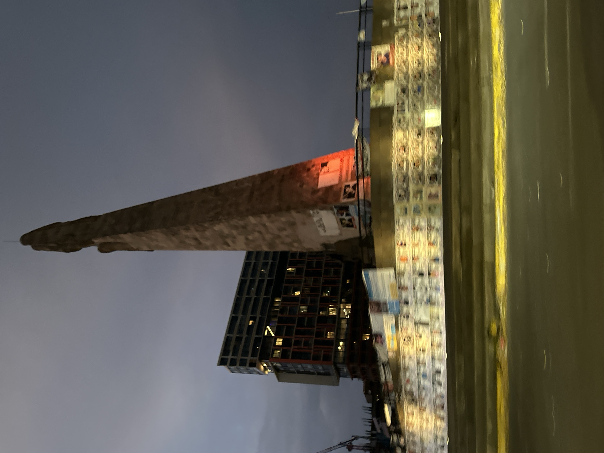 The image depicts an urban night scene in Guadalajara, Mexico, focusing on the iconic "Los Arcos" monument. The stone arch of Los Arcos stretches diagonally across the frame, with various posters and flyers affixed to its surface. Adjacent to the monument is a modern high-rise building with illuminated windows, suggesting residential or commercial occupancy. A street runs along the side, its surface reflecting ambient light, and a wall covered in posters stands near the road. The sky is a dark, muted blue, indicating nighttime. Construction equipment, specifically a crane, is visible in the lower right corner, suggesting nearby development activities. There are no people visible in the photograph.