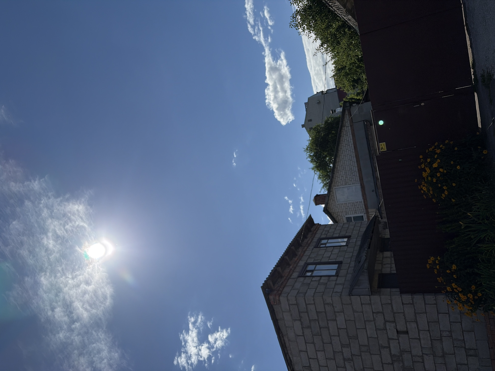 The image depicts a view of buildings under a bright, sunny sky with some wispy clouds. In the foreground, a dark brown fence stretches across the bottom half of the frame, with a cluster of yellow flowers and green foliage visible in front of it on the left. To the left of the fence is a partially constructed building made of gray cinder blocks, with two windows visible on its upper story. Behind this building and the fence, other structures are partially visible, including a smaller brick building with a chimney and a taller, modern-looking building in the distance. Green trees are interspersed among the buildings. The sun is high in the sky, creating a lens flare effect in the upper left quadrant of the image. The scene suggests an outdoor, possibly residential or industrial, area. The style of construction and the overall atmosphere are consistent with scenes found in various neighborhoods of Chernihiv, Ukraine, particularly in areas undergoing development or with older, single-family homes.