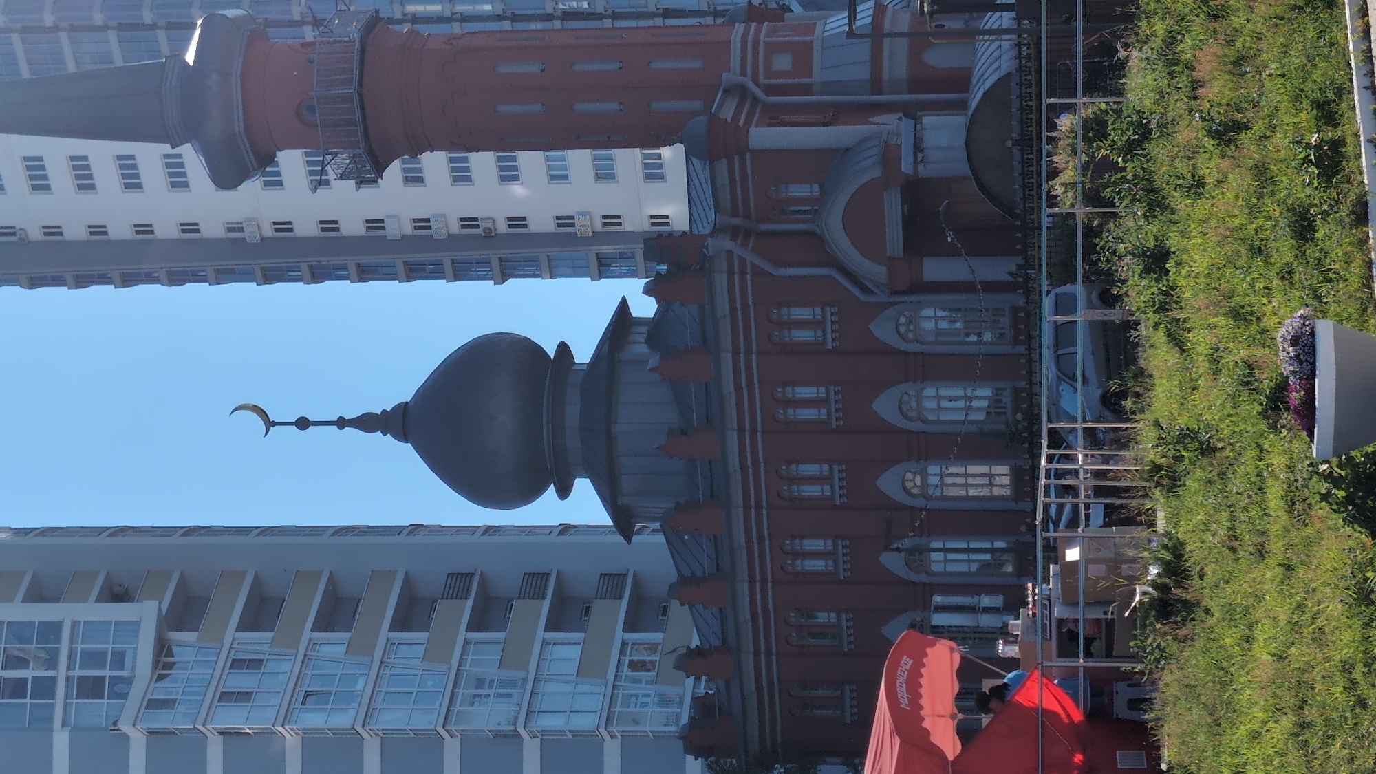 The image captures an outdoor scene with a striking architectural contrast in Nizhny Novgorod, Russia. The foreground features a vibrant red awning with Cyrillic text that reads "МАЙОКЕ" (Mayokye). Underneath the awning, two individuals are partially visible, suggesting a public space or event.
Dominating the background is a multi-story brick building with ornate detailing and a distinctive dark dome topped with a crescent moon. This architectural style hints at a historical or culturally significant structure, possibly a mosque or a building with a similar aesthetic influence. Adjacent to this, a tall, modern apartment building with a white facade and numerous windows stands, creating a juxtaposition of old and new architecture.
To the right of the buildings, a lush green grassy area with some vegetation and a large grey planter suggests a park or landscaped public space. A fence separates the grassy area from the buildings. The sky is a clear, bright blue, indicating fair weather and likely daytime, possibly mid-morning or afternoon. The overall impression is one of a city blending historical charm with modern development, set against a backdrop of clear skies and greenery.