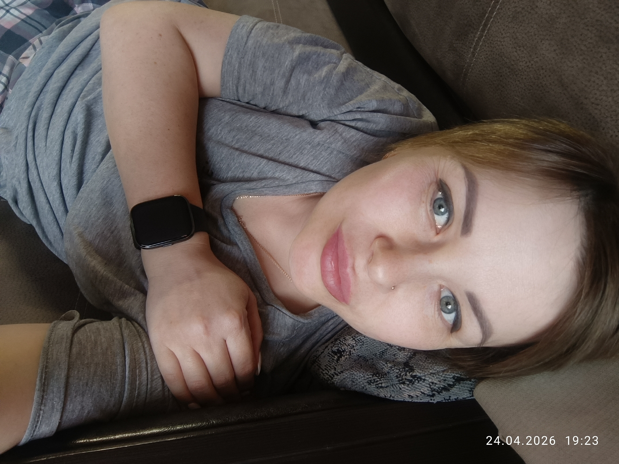 The image captures a woman lying in a relaxed pose on a brown sofa or couch, looking directly at the viewer. She is wearing a simple gray t-shirt, and a plaid-patterned fabric, possibly a blanket or pajamas, is visible to her left. A black smart watch is on her left wrist, and a thin, gold-colored necklace adorns her neck. She has light-colored skin, light blue-grey eyes, and short brown hair. Her facial features include natural-looking makeup, with subtle eyeliner and filled eyebrows, and her lips appear to have a pink tint. A small, silver-colored stud piercing is visible on her left nostril. Her expression is calm and engaging, with a slight hint of a smile. The setting is indoors, presumably a home in Chaikovsky, Russia. The date "24.04.2026" and time "19:23" (7:23 PM) are visible in the bottom right corner, indicating an evening shot.