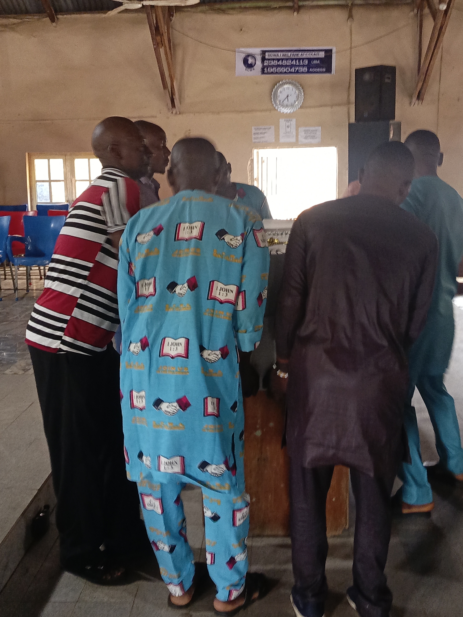 The image shows a group of men standing in an indoor space in Akwanga, Nigeria. A man on the left wears a striped shirt and black pants. Another man, centered in the frame, is wearing a blue outfit patterned with images of books and handshakes. There is a man wearing a dark colored outfit. In the background, blue chairs are visible. A sign is mounted on the wall. A clock hangs above some paper or documents that are also on the wall. A speaker is in the top right corner. The indoor space appears to be a simple room.