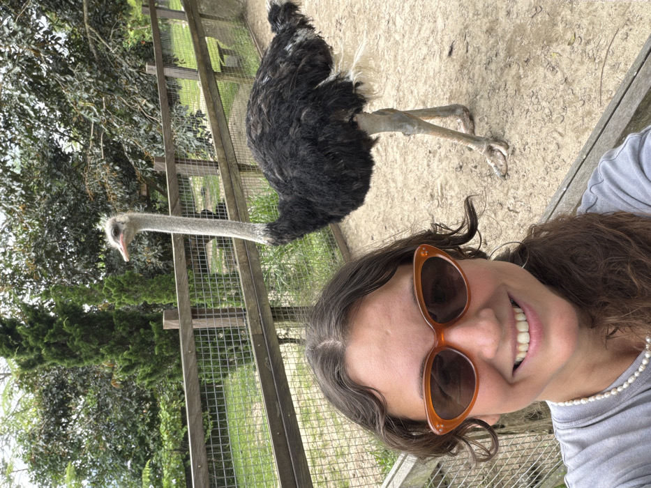 A woman wearing sunglasses poses for a selfie. In the background, a black and gray ostrich stands behind a wooden fence with a wire mesh. Green foliage is visible behind the fence on the left. The ground behind the ostrich is dirt. The woman is smiling, and she is wearing a pearl necklace.