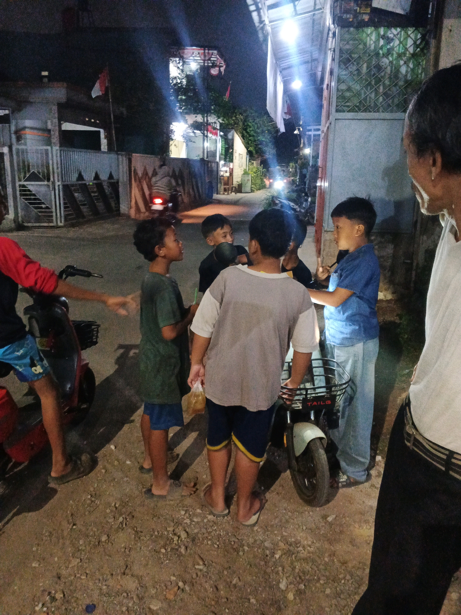 In Kadu, Indonesia, at night, a group of four young boys and an older man are gathered around a small electric scooter on a dirt road. The boys appear to be engaged in conversation or play. One boy in a green t-shirt is holding a small plastic bag. Another boy in a blue shirt and jeans is holding his hand up. The older man, seen from the side, has dark hair and is wearing a white shirt. The environment is dimly lit, with overhead lights casting shadows and illuminating the scene. Behind the group, a fence and a building are visible, suggesting a residential area. The scooter has a basket on the front with the word "TAILS" visible.