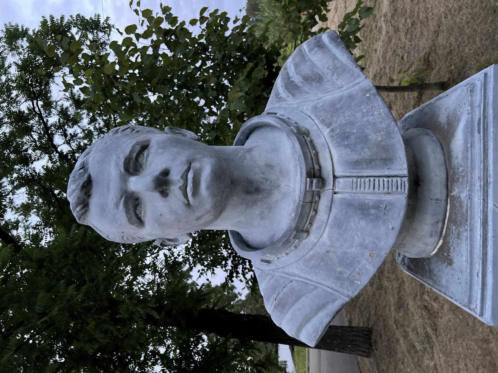 A gray bust of a man in a spacesuit stands on a pedestal outdoors. The bust depicts a man with short, combed-back hair, a serious expression, and a raised collar typical of an astronaut's suit. The statue is weathered, with visible cracks and peeling paint. Behind the bust, lush green trees and a cloudy sky form the backdrop. The ground in front of the pedestal is covered in dry grass and straw. The location is the Yuri Gagarin Park in Krasnodar, Russia.