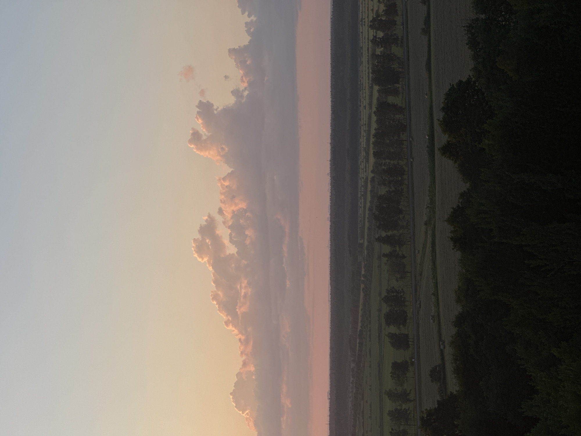 An aerial view captures a rural landscape at dusk. The sky transitions from a soft blue at the top to a pale pink and peach hue near the horizon, illuminated by the setting sun. Cumulus clouds, tinged with pink and orange, drift across the sky. Below the sky, a dark, distant treeline forms a horizontal band across the scene, marking the transition to the land. The middle ground features a patchwork of fields, some appearing as agricultural land and others as meadows, with a line of trees running along a road or pathway. Several round hay bales are visible in the fields. In the foreground, a dense cluster of dark green trees fills the bottom of the frame, obscuring the ground beneath them. The overall impression is a serene, natural setting at the close of day.