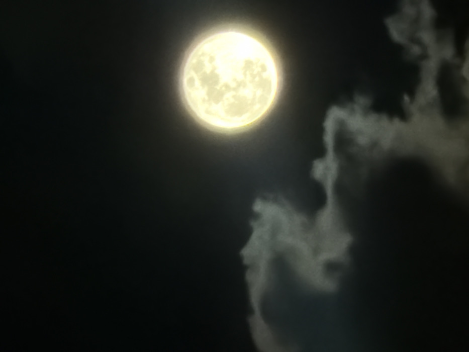 The image depicts a nocturnal sky scene over Tel-Aviv, Israel. A brightly illuminated, full moon, appearing as a yellow-white sphere with faint surface characteristics, is positioned in the upper right quadrant of the frame. Below and to the left of the moon, dark, layered cloud formations are visible, partially obscuring the deep black background sky. No persons, man-made objects, or ground-level features are discernible. No actions or interactions are depicted within the captured scene.