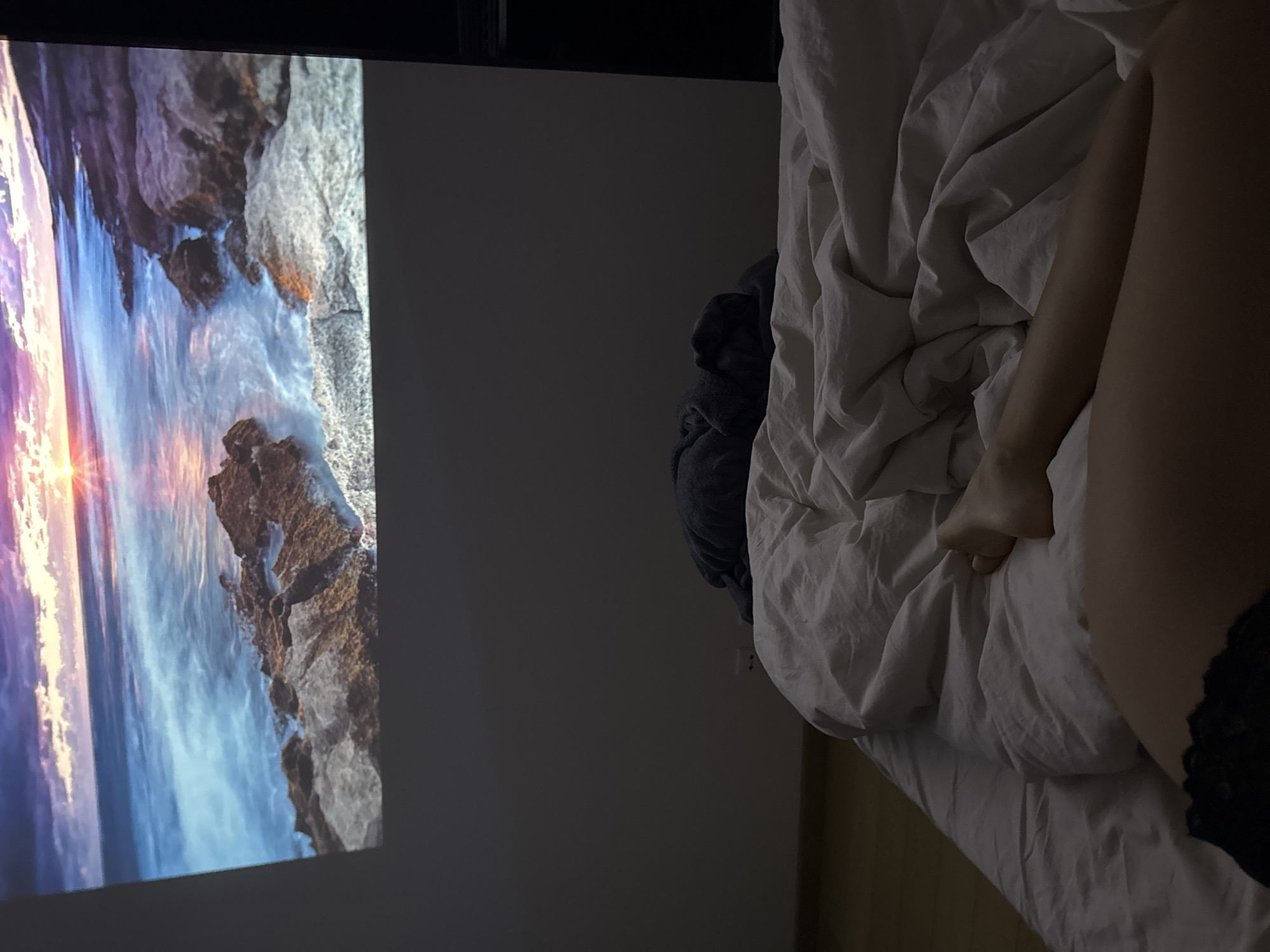 The image shows a dimly lit bedroom setting. On the left, a projected image of a coastal scene with rocks, water, and a sunset is visible on the wall. To the right, a bed with crumpled white sheets is present. A person's leg and foot are visible on the bed, with black lace trim on their garment. No specific landmarks indicate the location being Warsaw, Poland.