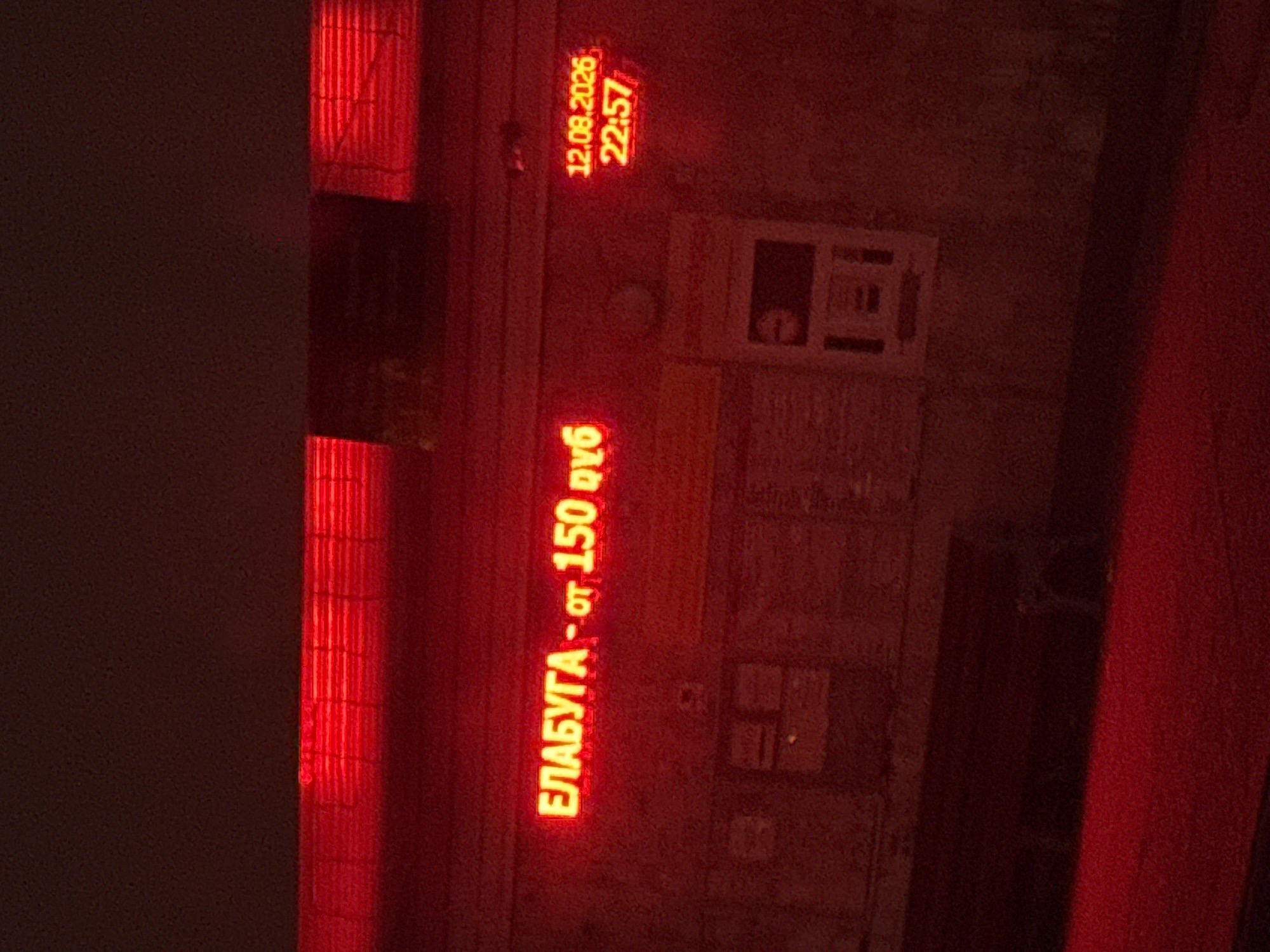 A nighttime exterior shot depicts a red neon sign displaying Cyrillic text and numerical information against a dark, textured building facade. The sign reads "ЕЛABУГA - от 150 ру6" in bright red letters, suggesting a price or service related to "Elabuga." To the right of this text, a digital display shows the date "12.08.2026" and the time "22:57" in red digits. The building appears to have a series of horizontal ribbed panels above the neon sign. Below the sign, there are some indistinct rectangular objects, possibly informational displays or windows. The overall impression is that of a commercial establishment or information kiosk, illuminated by its own signage. The location is identified as Oktyabr'-Bulyak, Russia.