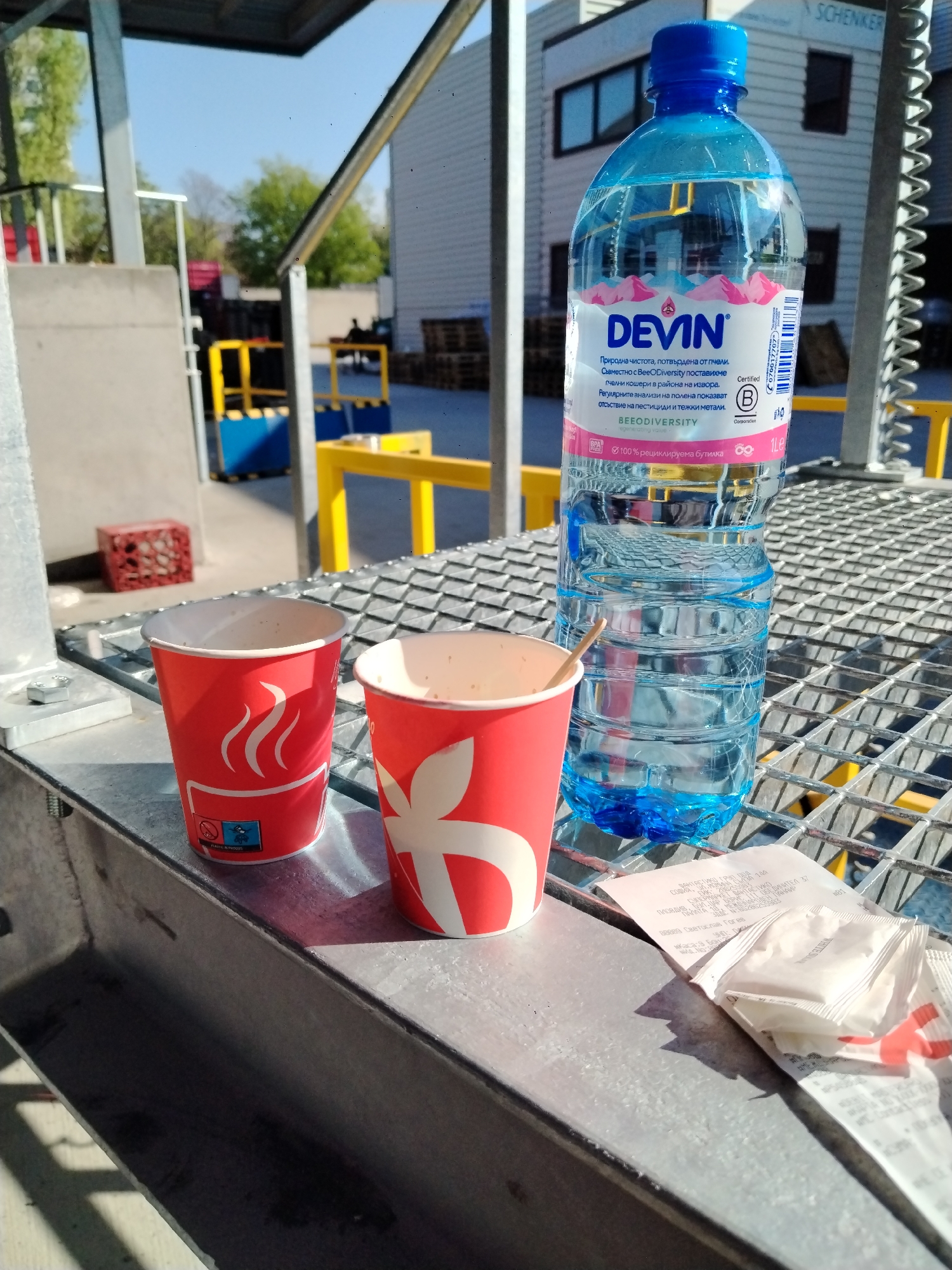 The image shows two red paper coffee cups, a bottle of Devin water, a receipt, and sugar packets on a metal grate surface. The receipt includes the name "ОБЕДИНИТЕЛ," an address on "БУЛ. ЦАР БОРИС III ОБЕДИН," and "ПЛОВДИВ" indicating the location is in Plovdiv, Bulgaria. In the background, there is a building with the word "SCHENKER" visible. There is also some yellow railing and a metal structure visible in the background. The environment appears to be industrial or commercial outdoor setting. There are no people visible in the image.