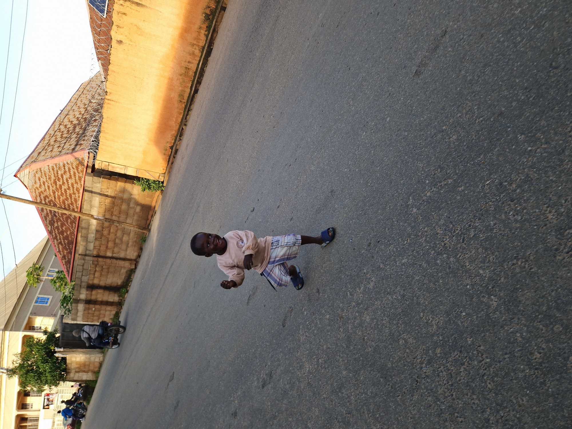 A young Black boy is captured in motion on a paved, sloping road. He is wearing a long-sleeved light brown shirt with a circular emblem on the left chest and plaid shorts. The boy appears to be mid-stride, with his right leg forward and his left arm raised, and his mouth open in a joyful expression. He is wearing dark blue sandals. In the background, a wall of cinder blocks forms the side of a building. Beyond the wall, a sloping roof of corrugated metal is visible, casting shadows on the wall. Another building with a light-colored facade and blue-framed windows is visible further in the distance. To the left of the frame, a motorcycle is parked, and another motorcycle is seen in motion with a rider. The road is mostly empty except for the boy and the distant vehicles. The lighting suggests it is late afternoon, with the sun casting long shadows.
