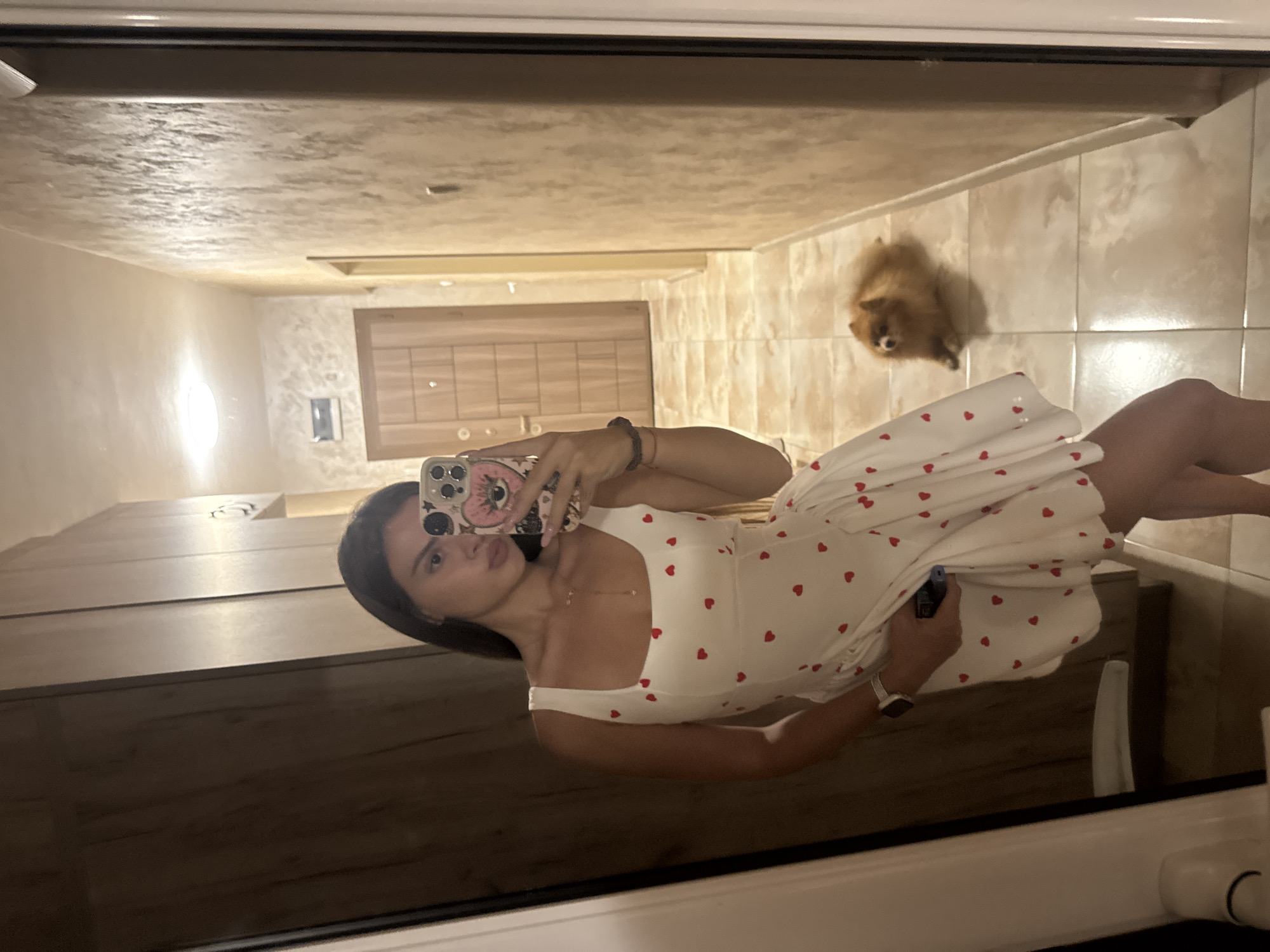 This image is a mirror selfie taken by a female individual within the interior hallway of a residence located in Plovdiv, Bulgaria.
The female subject, centered in the frame, has long, dark, straight hair, a medium skin tone, and is wearing makeup. She is dressed in a white sleeveless mini-dress adorned with numerous small red heart patterns. The dress features a square neckline and a pleated skirt. On her left wrist, she wears a silver-colored watch, and on her right wrist, she has multiple dark-colored bracelets. A delicate gold-colored necklace with a small pendant is visible around her neck. She holds a smartphone with a distinctive pink case featuring an eye design in her right hand, positioned to capture her reflection. Her left hand is partially obscured, holding a small dark object (possibly another phone or remote) tucked near her hip. She maintains a serious or slightly pensive expression, looking towards the camera's reflection.
Further down the tiled hallway, a small, fluffy dog, appearing to be a light brown/orange Pomeranian, is lying on the floor.
The environment consists of a well-lit hallway with light-colored, square-tiled flooring that has subtle mottled patterns. The wall reflected on the left side of the image features dark brown wood paneling, suggestive of a built-in closet or wardrobe. The wall reflected on the right side has a light beige/tan textured finish. A circular, flush-mounted light fixture illuminates the white ceiling. In the background, two wooden interior doors are visible; one on the left has a modern design with horizontal panels and a silver handle, while another doorframe is partially visible further down the hallway on the right. A small rectangular wall-mounted device (possibly an intercom or thermostat) is visible above the left door. White baseboards line the bottom of the walls. The mirror itself has a visible white frame on the left side.