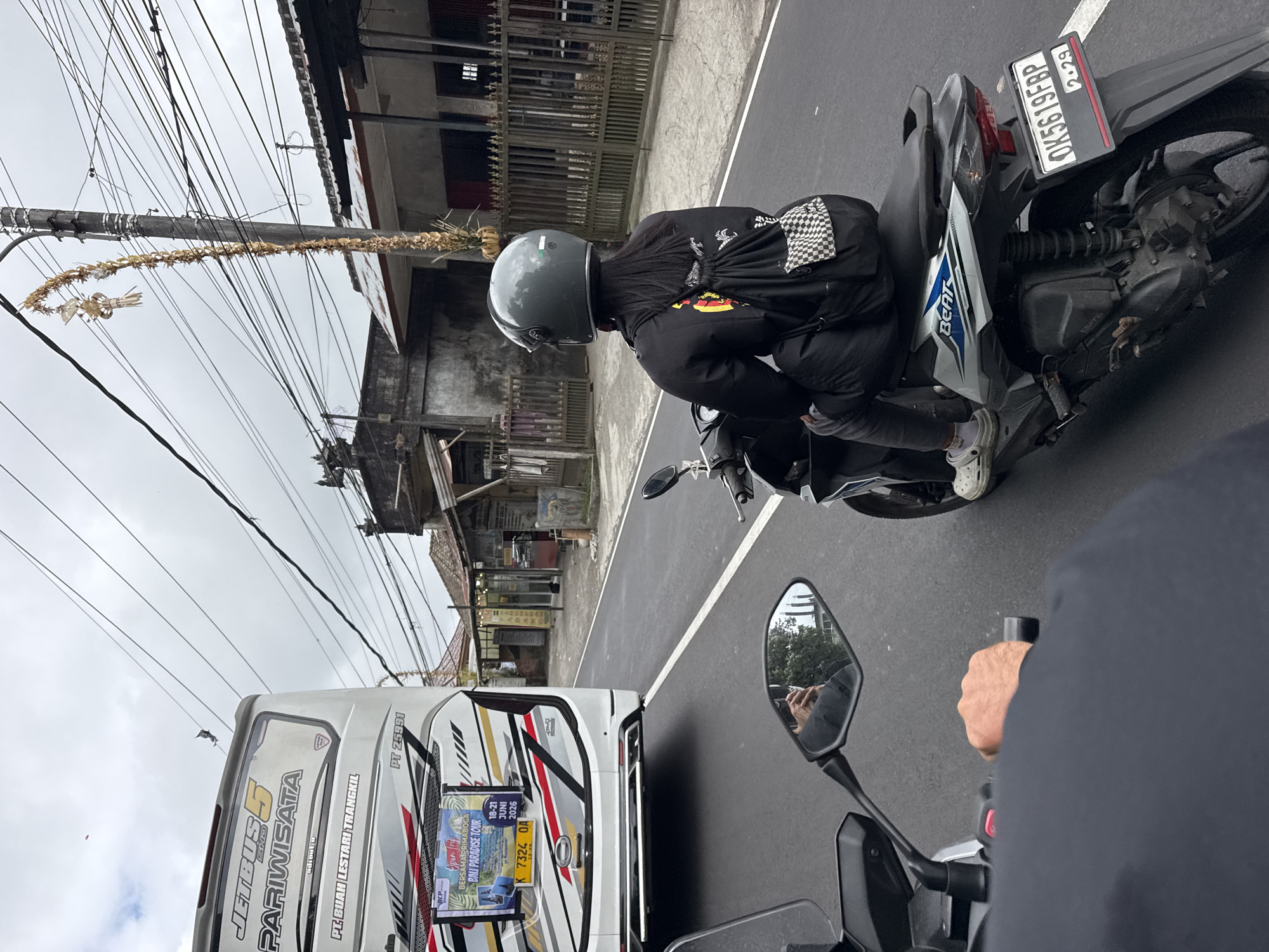 The image depicts a street scene in Kintamani, Indonesia, with a focus on two-wheeled vehicles and a bus. In the foreground, the rider's perspective of a motorcycle is visible, showing a hand on the handlebar. To the right, another motorcycle is being ridden by a person wearing a grey helmet and a black jacket with a checkered backpack. The motorcycle has a license plate reading "OK5619FBP". To the left, the rear of a white "Jetbus 5 Pariwisata" bus is seen, featuring advertisements and license plate "K 7324 OA". The environment shows a paved road with white lane markings, lined by buildings with tiled roofs and a wooden fence. Overhead, a dense network of electrical wires crisscrosses the sky, which is overcast. A traditional Balinese "penjor" (a tall, curved bamboo pole decorated with offerings) is visible, suspended from a utility pole.