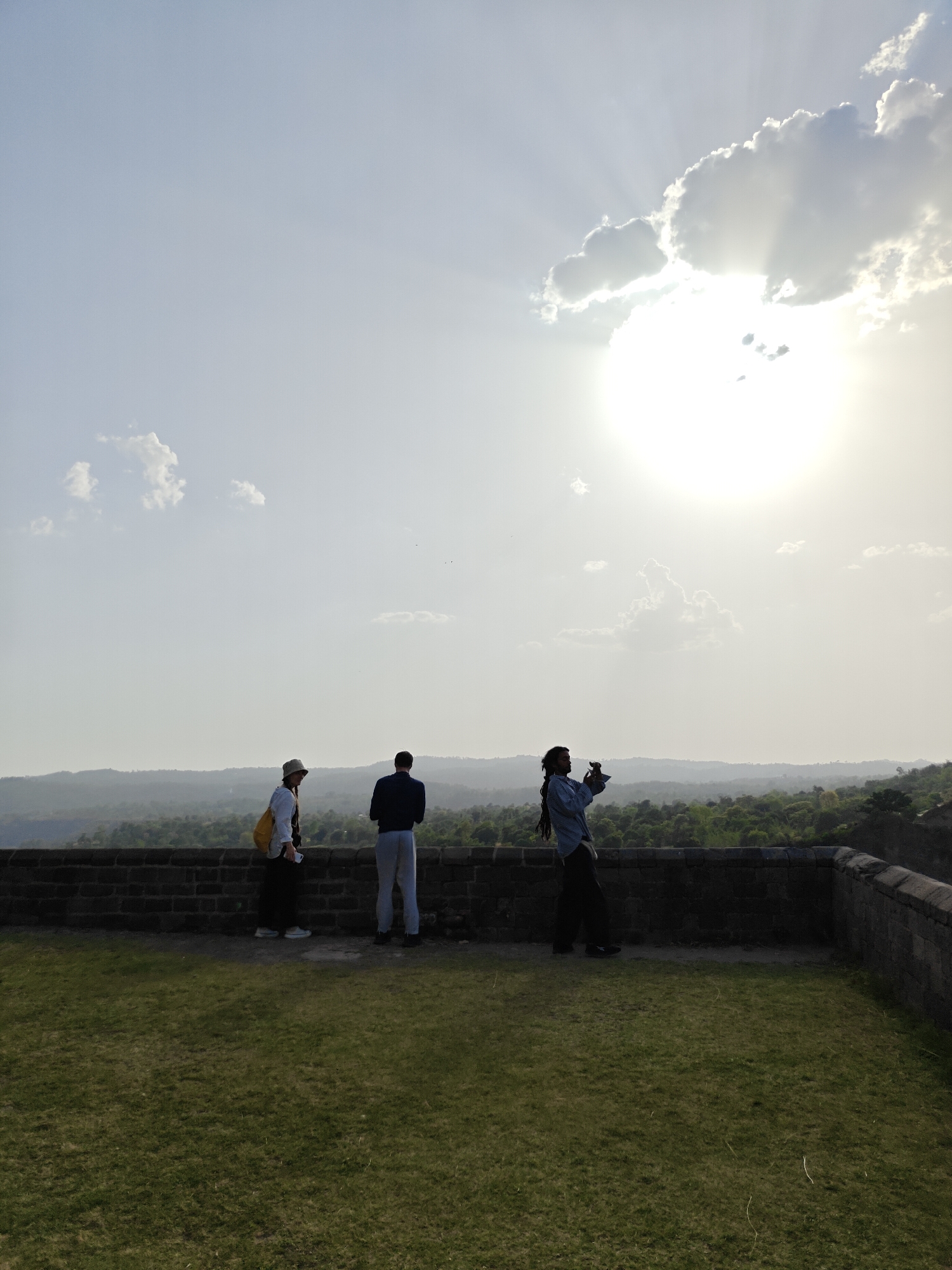 Three individuals stand on a brick parapet overlooking a wooded landscape in Kaswara, India. The person on the left wears a hat and carries a yellow bag. The middle person faces away from the camera, wearing a dark jacket and light-colored pants. The person on the right appears to be taking a picture. Above, the sun shines brightly through clouds, creating a hazy sky. Grass covers the ground in the foreground.