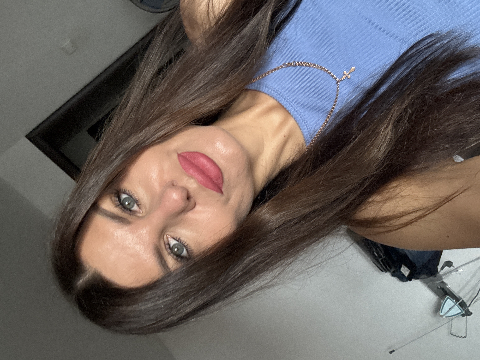 The media file is a close-up, vertically oriented selfie of a woman. She has long, dark brown hair that partially obscures her face and shoulders. Her eyes are a striking green and her lips are painted with a bold pink lipstick. She is wearing a light blue, ribbed top and a delicate gold chain with a small cross pendant. The background appears to be an indoor setting with a plain wall and a dark horizontal element that could be a window or a piece of furniture. The lighting suggests it is daytime. There is no visible text in the image.