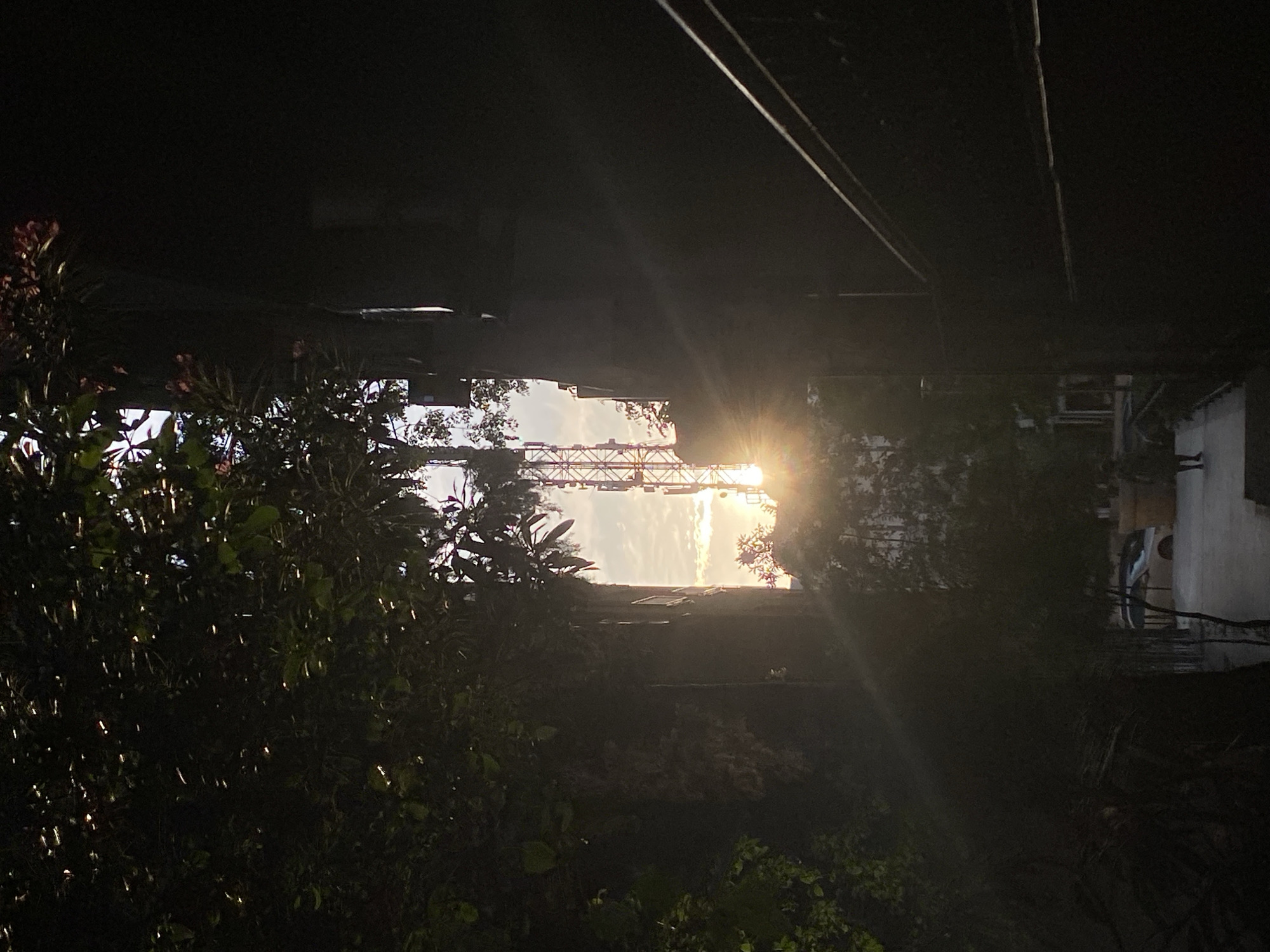 The photo is an upward-looking view in a built environment in Podgorica, Montenegro. The sun is prominently visible in the central portion of the image, partly obstructed by a metal structure similar to a bridge or utility tower. Above, dark structures could be parts of buildings or infrastructure. Vegetation, including trees and plants, is visible at the bottom of the frame and on the left. On the right side, there are parts of a building, a balcony, and powerlines. The overall lighting suggests it is either sunrise or sunset.