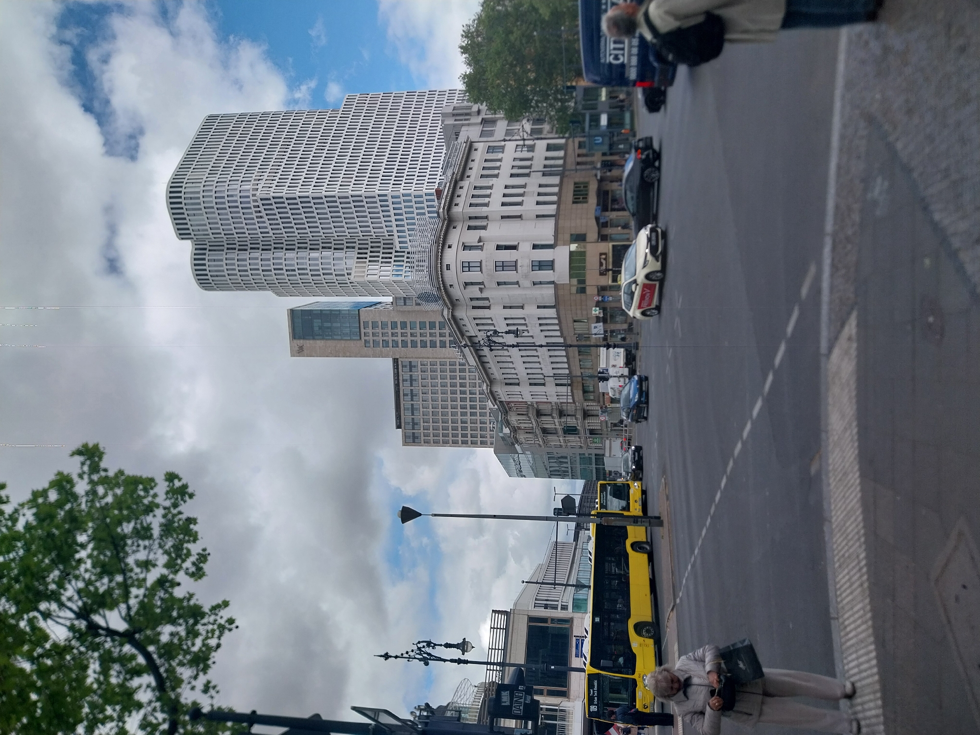 The image shows a street scene in Berlin, Germany. Prominently visible is the Upper West building complex, a modern architectural structure. A multi-story building with a grid-like facade stands adjacent to it.
A city street runs along the base of the buildings, where vehicles, including a yellow bus and several cars, are visible. Pedestrians are present along the sidewalk. One individual is walking on the right side of the frame, while another is standing near the bus stop on the lower portion of the frame.
Lampposts are visible along the sidewalk. The sky is filled with a mix of clouds and patches of blue sky.