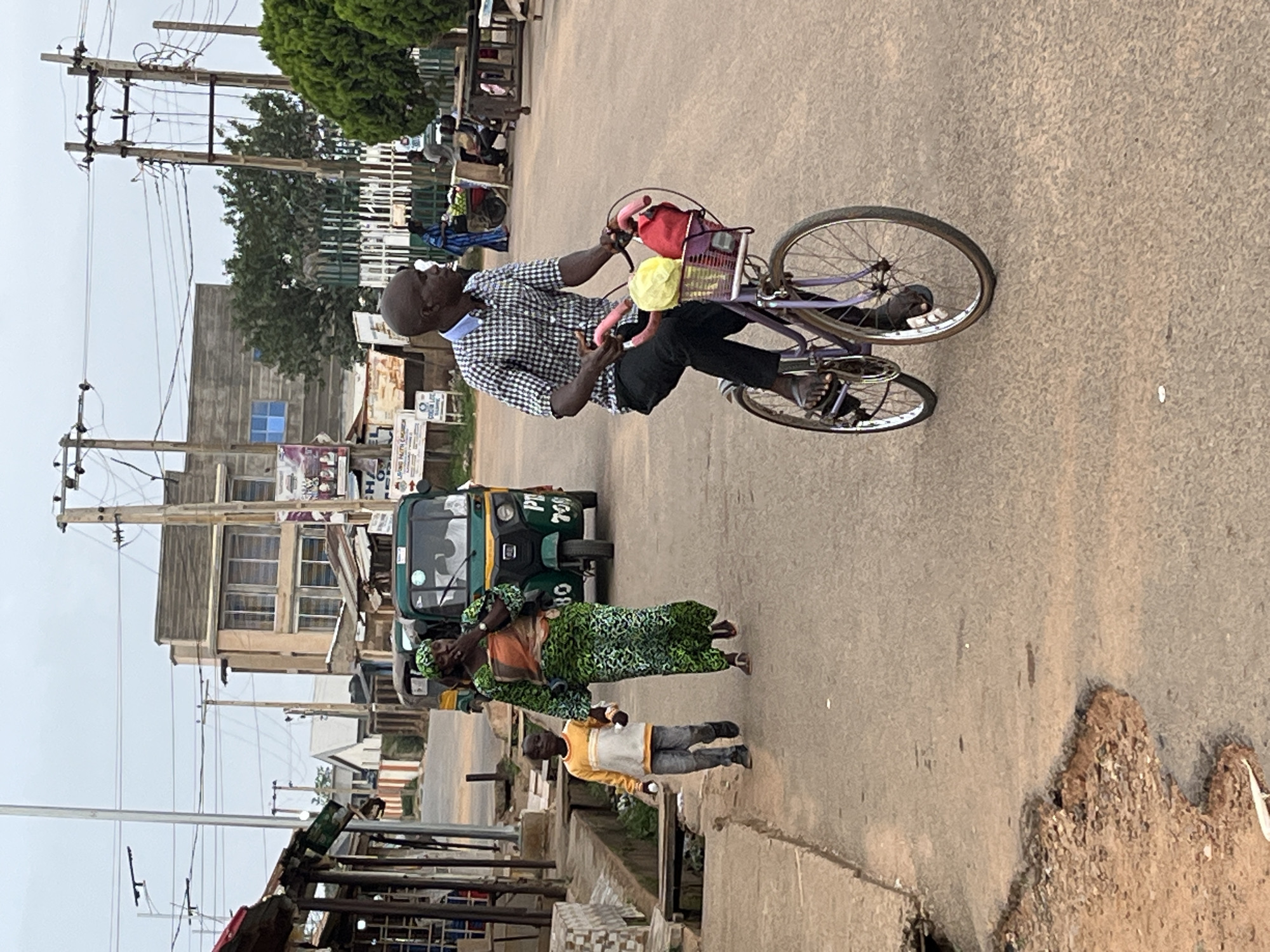 The image depicts a street scene in Barakin Akawo, Nigeria. A man rides a purple bicycle along a paved road. The bicycle has a front basket containing what appears to be a red bag and a yellow object. The man wears a checkered shirt and black pants. A green auto-rickshaw is parked nearby. Two adults and a child are walking along the sidewalk. Overhead are numerous utility poles with wires. Buildings are visible in the background. The road surface shows signs of wear.