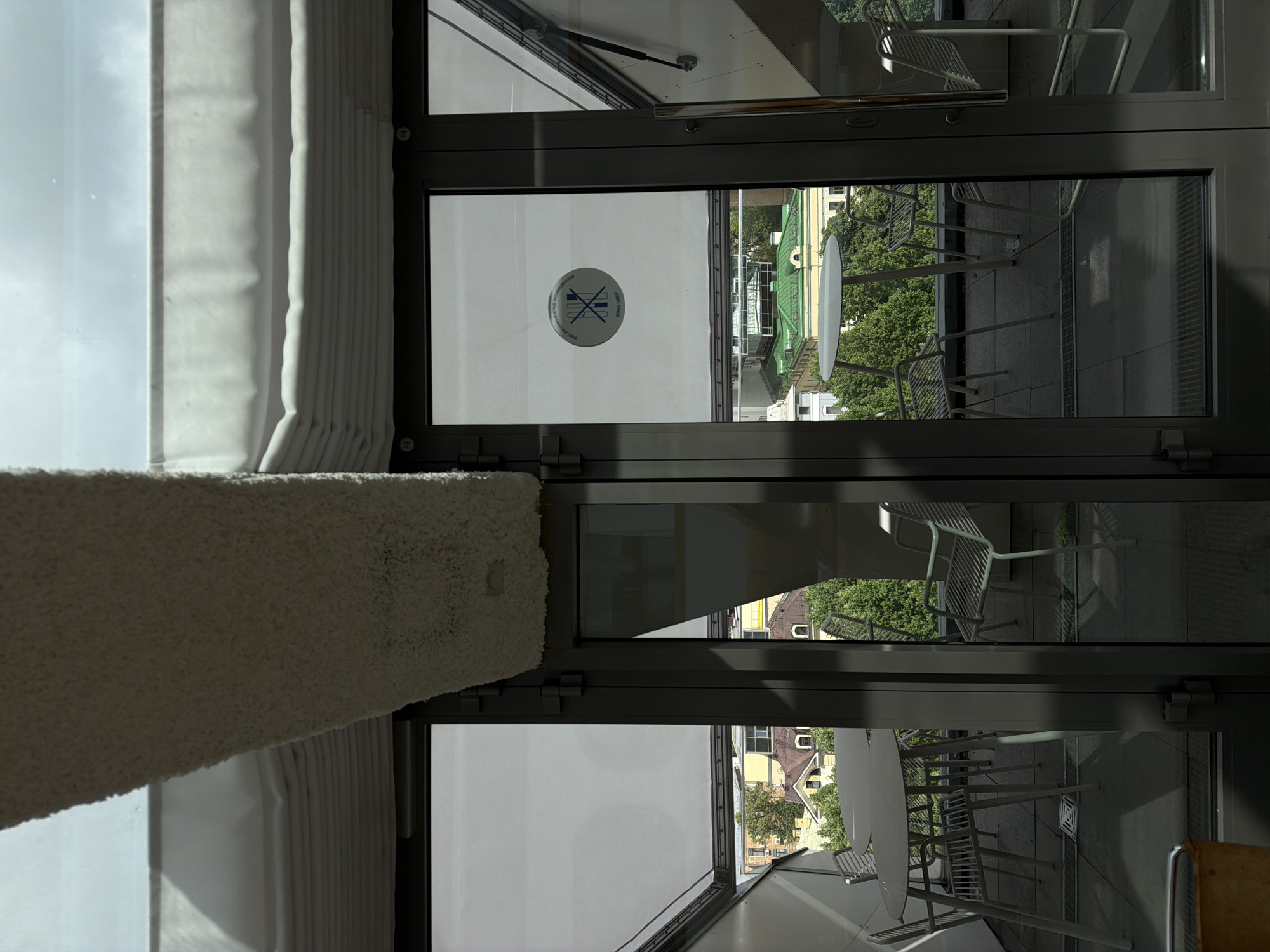 The scene shows a view through a window, possibly in Moscow, Russia, based on architectural styles and potential signage visible in the reflection. The window is divided into sections, with dark grey metal frames.
Visible outside the window is a balcony or patio area with outdoor furniture, including white tables and chairs. Buildings and trees are visible beyond the balcony, suggesting an urban setting.
A sticker or sign is affixed to the window glass, with a logo.
Curtains or blinds are partially visible in the immediate foreground.
The photograph is taken from an interior perspective looking outward.