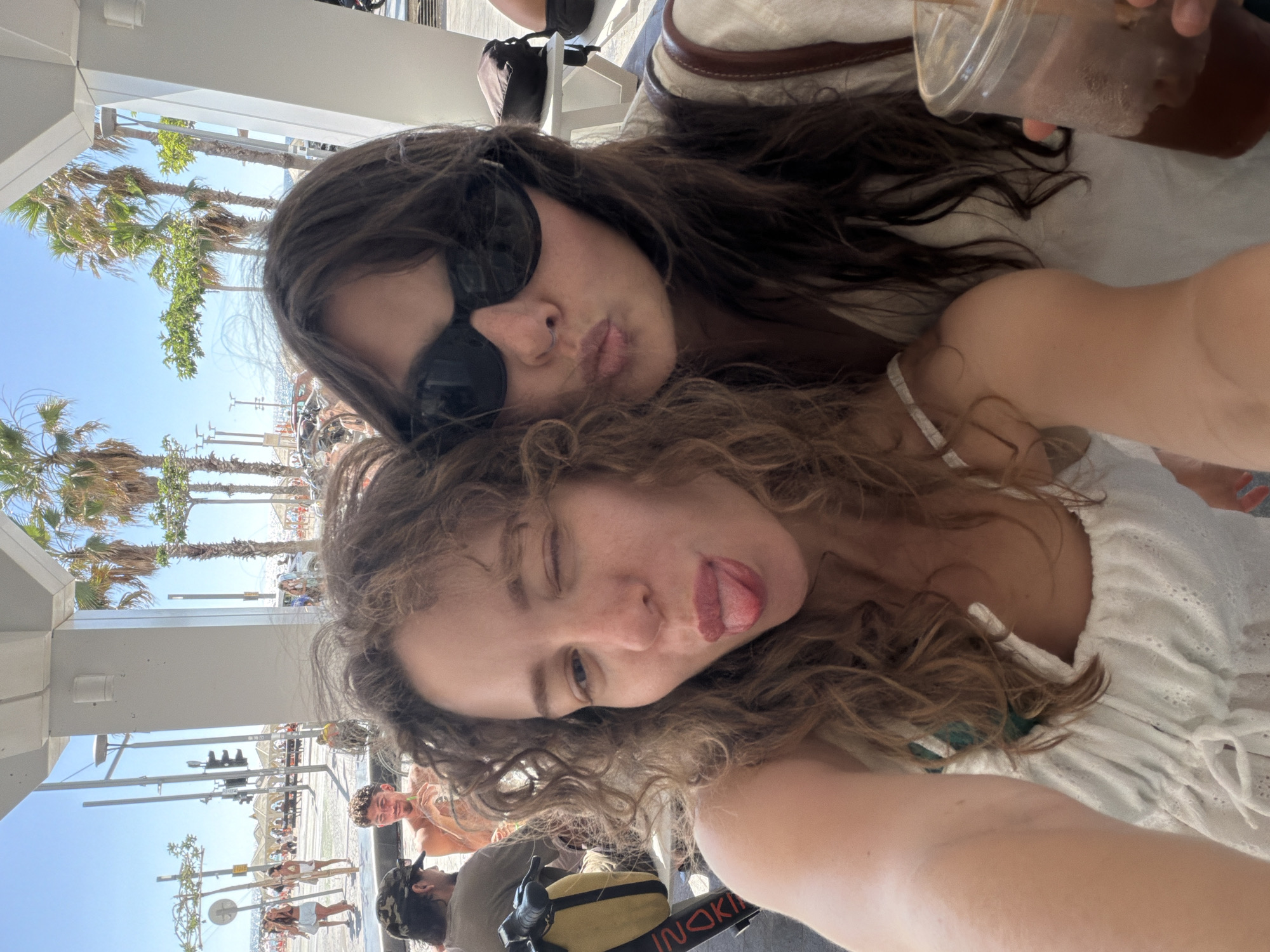 This media file depicts two young women at a beachside location, likely in Tel Aviv, Israel, on a bright, sunny day. The foreground is dominated by the faces of the two women. The woman in the upper part of the frame has dark hair and is wearing sunglasses, with her lips pursed as if blowing a kiss. The woman below her has curly, light brown hair and is winking, with her tongue sticking out. She appears to be wearing a white, eyelet-detailed top.
In the background, there are glimpses of a bustling beach scene. Palm trees are visible, along with beach umbrellas and people lounging on the sand. Beyond the beach, there's a clear blue sky. Structures, possibly part of a boardwalk or pier, are also present, with railings and overhead coverings suggesting a sheltered area. People are walking around, and there's a traffic light visible on a pole, indicating a public area with pedestrian and possibly vehicular traffic.
One of the women is holding a plastic cup with a beverage, suggesting they are enjoying drinks. The overall mood appears to be playful and relaxed, indicative of a holiday or leisure outing. The bright sunlight and beach setting clearly point to daytime. There is some visible text on a black strap in the lower right corner which appears to read "INOKI".