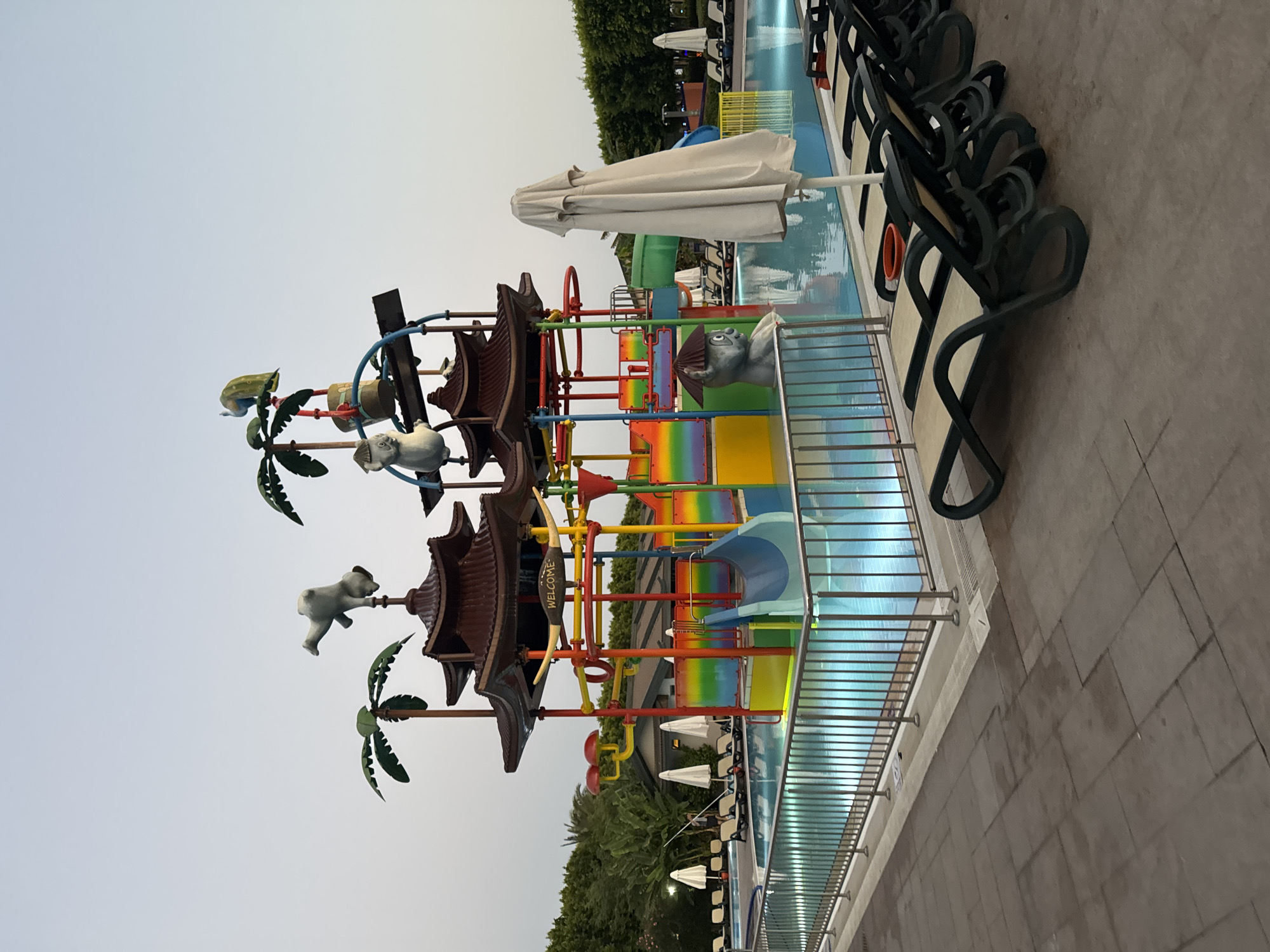 The image shows a water park complex at the Akka Antedon Hotel in Aksu, Turkey, during the twilight hours. The central feature is a multi-tiered water play structure with a pagoda-style roof, adorned with colorful slides, animal figures, and palm tree decorations. The structure is surrounded by a bright blue swimming pool, illuminated from beneath, which reflects the sky and surrounding elements.
In the foreground, a row of empty, dark green lounge chairs with beige cushions are neatly arranged on a tiled patio. To the right, a closed, light-colored umbrella stands upright. The pool is bordered by a silver metal fence. In the background, lush green trees and other hotel buildings are visible. The sky is a gradient of pale blue and grey, indicating the transition from day to night. There are no people actively engaged in the water park activities; the scene appears to be before the park's busy hours or after closing. The overall impression is one of tranquility and readiness for activity.