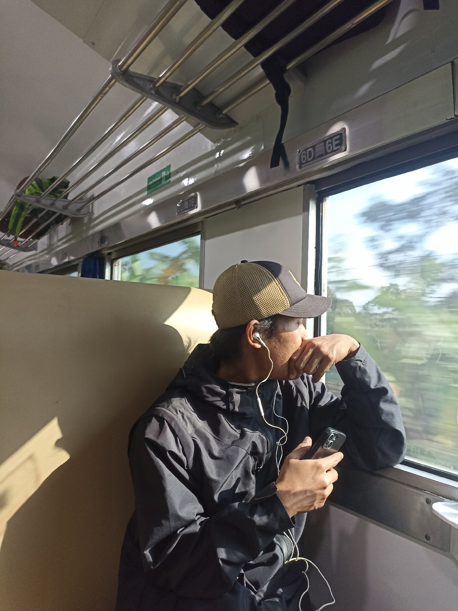 In the city of Sempu, Indonesia, a young man wearing a black jacket, a tan and black baseball cap, and white earbuds sits on a tan-colored seat inside a train carriage, looking out the window. He is holding a black smartphone in his right hand. Sunlight streams through the window, casting a shadow on the wall behind him. The train appears to be in motion, as indicated by the blurred scenery outside the window. Luggage is visible on a rack above the seats.