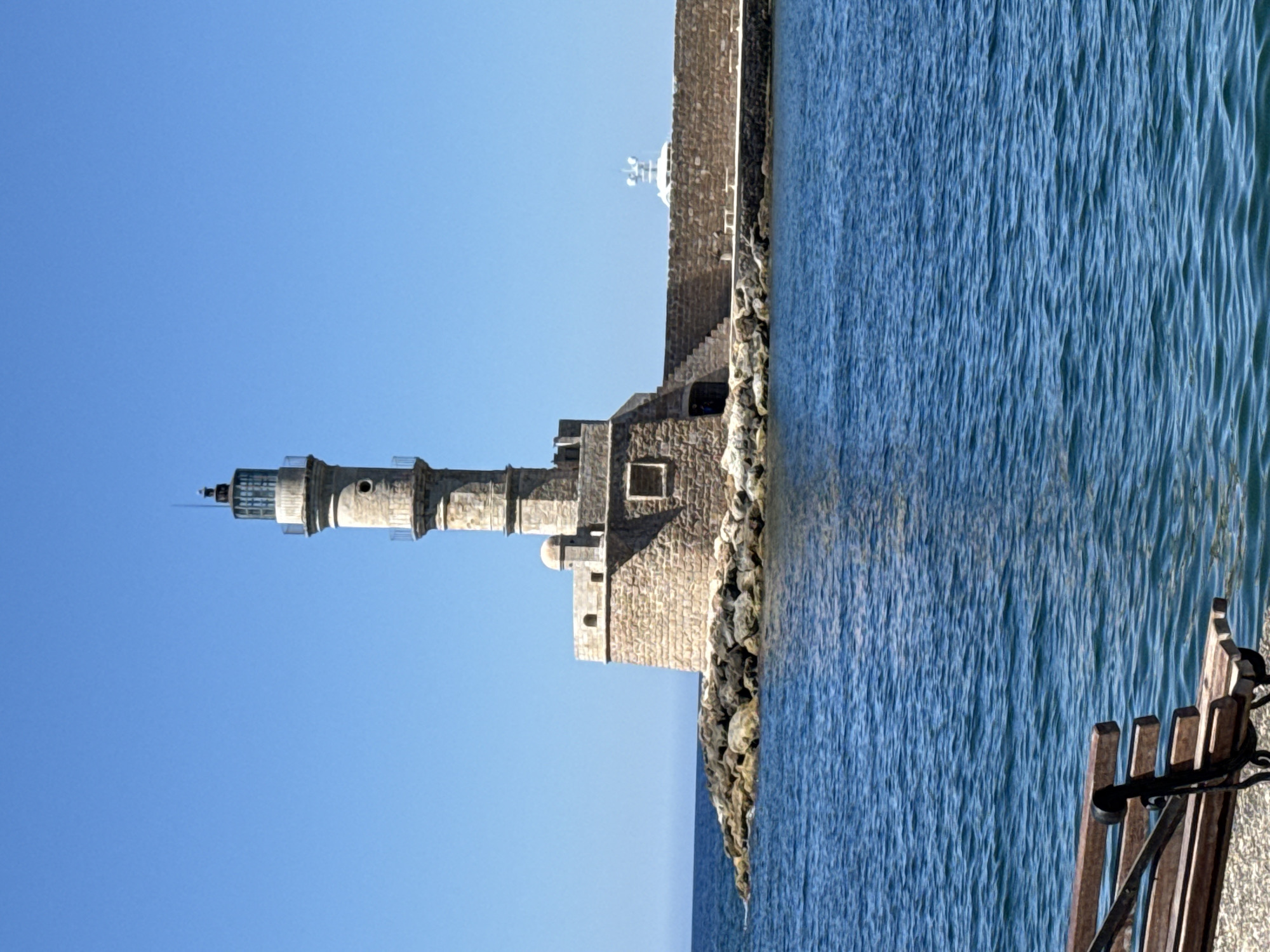 A wide, low-angle shot depicts the historic lighthouse of Chania, Greece, standing tall on a rocky outcrop. The lighthouse, constructed of weathered stone, has a cylindrical tower topped with a lantern room. It is situated on a mole extending into the bright blue sea, with a stone seawall visible to its right. The water is a deep, calm blue with gentle ripples. In the foreground, a wooden park bench with metal legs sits on a paved surface, facing the water and the distant lighthouse. The sky above is a clear, cloudless blue. A white structure, possibly a navigational aid, is visible on the seawall further down. The overall scene is serene and captures the iconic landmark under a sunny sky.