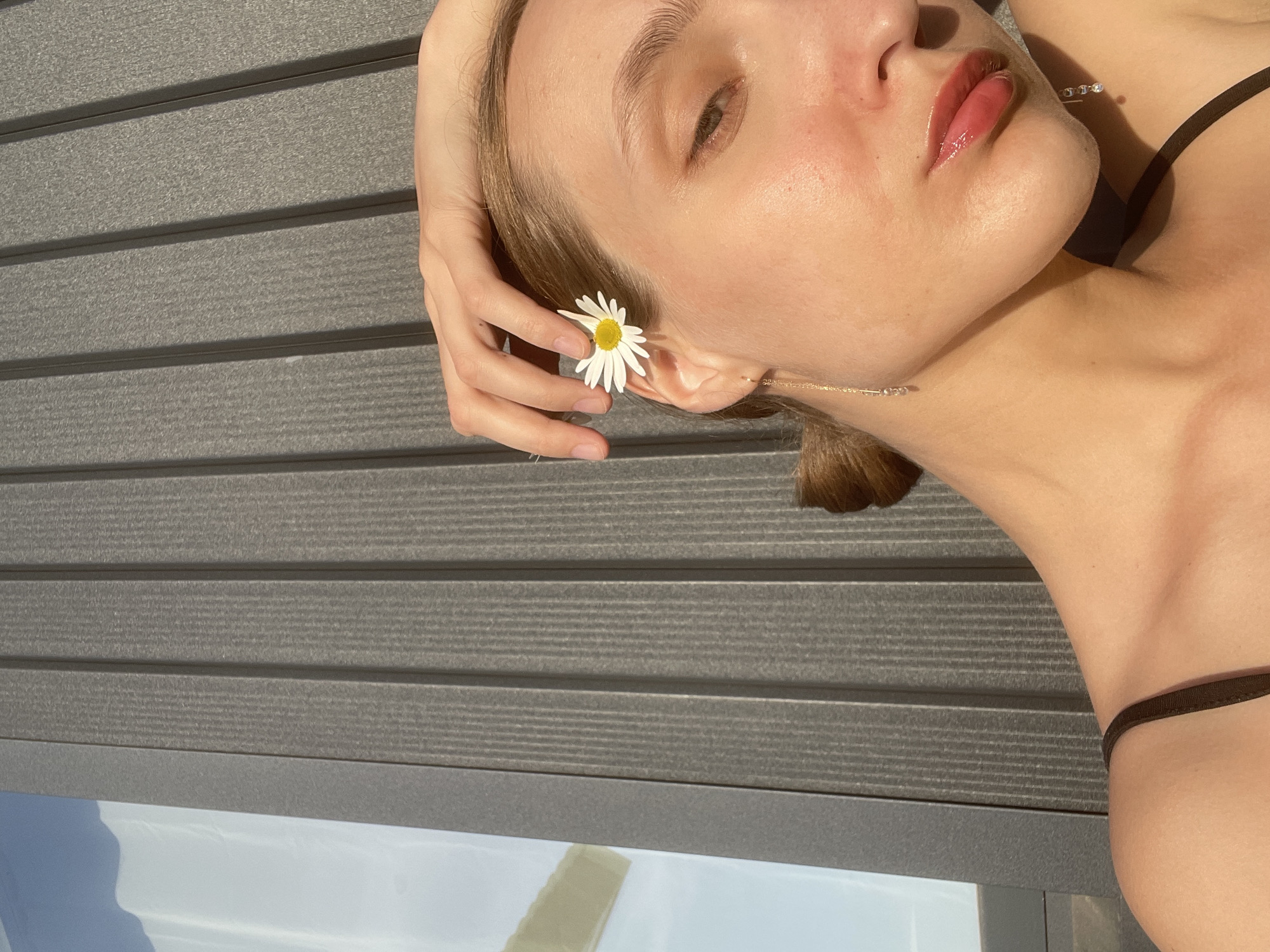 The image shows a woman lying down outdoors. She has fair skin, light brown hair pulled back, and wears a thin black strap indicating a garment like a bikini top or tank top. She is holding a daisy flower next to her ear. A delicate gold chain necklace is visible on her neck. The background consists of gray horizontal planks, possibly decking or siding. A reflection in the lower left corner hints at a nearby reflective surface, potentially water or glass. There is no indication that this image was taken in Подшивалово, Russia.