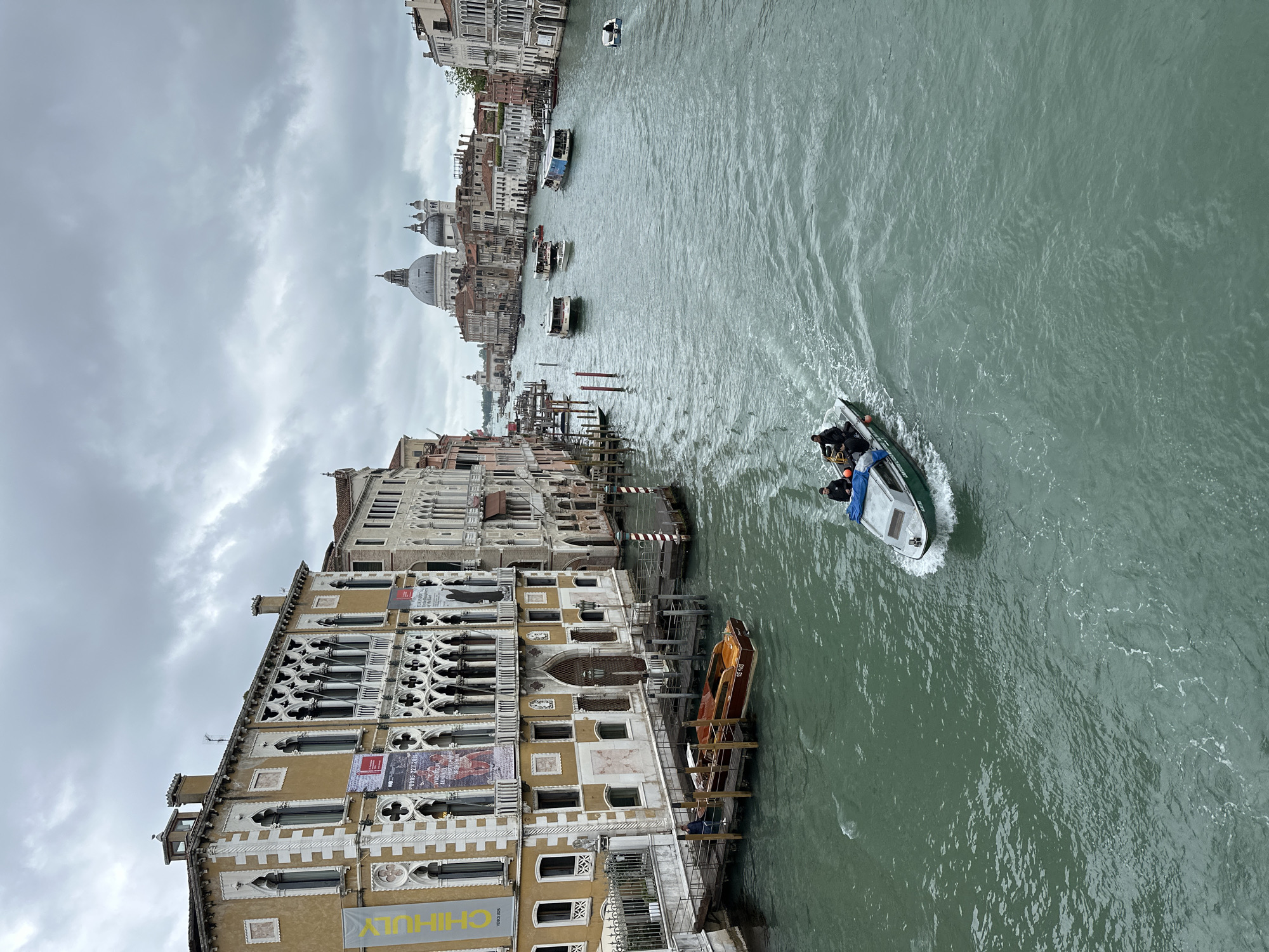 The image presents a view of Venice, Italy, specifically capturing a section of the Grand Canal. The perspective is from an elevated position, likely a bridge or high building.
On the left side of the frame are Venetian buildings characterized by unique architecture, including intricate latticework, arched windows, and textured facades. One building displays posters or banners on its exterior.
In the center of the frame, the Grand Canal stretches into the distance, reflecting the cloudy sky above. Several boats populate the waterway, including a water taxi moving through the canal, creating waves. A few other boats are docked.
In the background, the iconic dome of the Santa Maria della Salute church is visible. The sky is overcast, contributing to the atmospheric quality of the image. The water has a greenish hue. There are at least two individuals in the boat in the water.