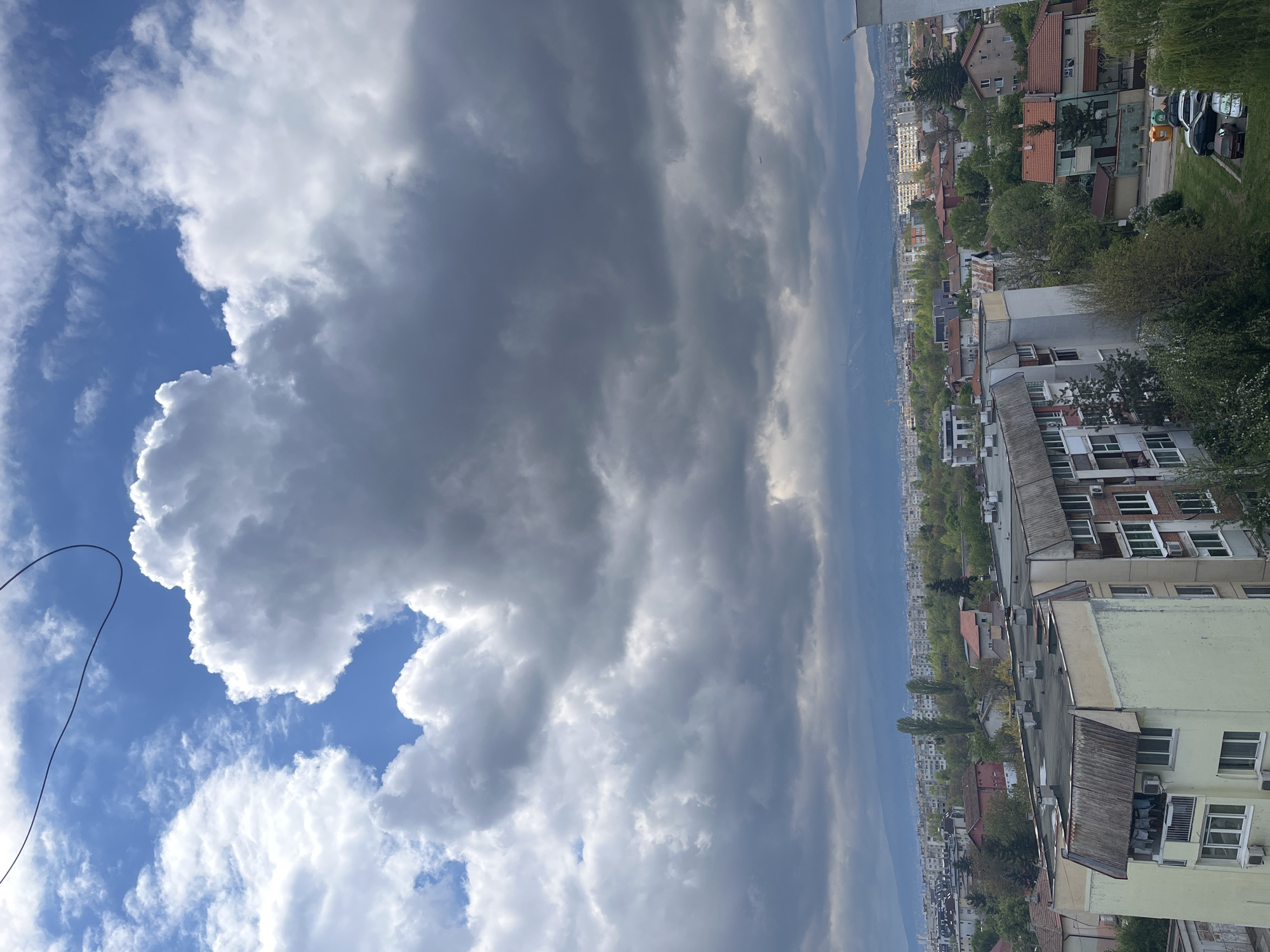 A sideways view from a high vantage point shows part of Sofia, Bulgaria, on a cloudy day. The sky, predominantly cloudy with sections of bright blue, takes up the majority of the view. To the right, residential buildings, various homes and structures dot the landscape, interspersed with trees. In the background, indistinct hills or mountains are visible in the distance, enveloped in haze.
