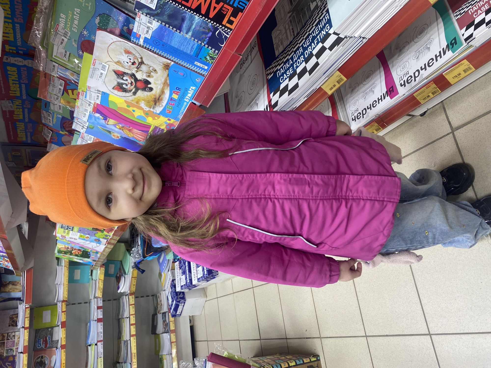 A young girl stands smiling in the aisle of what appears to be a stationery or art supply store. She is positioned slightly off-center to the left, looking directly at the camera. The girl wears a bright pink puffer jacket, an orange knit beanie with "AYC STREET" written on it, blue jeans, and dark shoes. Her long brown hair flows over her shoulders.
The background is filled with shelves stocked with various paper products and art supplies. On the left, products like "ЦВЕТНАЯ БУМАГА" (Colored Paper) and "МЕЛОВАННАЯ БУМАГА" (Coated Paper) are prominently displayed, with price tags showing values such as "133.00", "258.00", and "79.00" rubles. Some paper packages specify "ДВУСТОРОННАЯ БУМАГА" (Double-sided Paper) and counts like "5 листов" (5 sheets) or "48 листов" (48 sheets).
On the right, taller shelves hold larger items, including folders and cases for drafting. Text like "ПАПКА ДЛЯ ЧЕРЧЕНИЯ" (Case for Drafting) and its English translation "CASE FOR DRAFTING" are clearly visible, along with "для черчения" (for drafting). These items also have price tags, such as "300.00", "178.00", and "157.00" rubles. The store has a tiled floor. The girl's warm clothing, including a jacket and beanie, suggests it might be cold weather outside, even though she is indoors. Her happy expression indicates she is enjoying her time in the store, possibly browsing for supplies. The prices and Cyrillic text confirm the location as Russia, aligning with the provided context of Izhevsk.