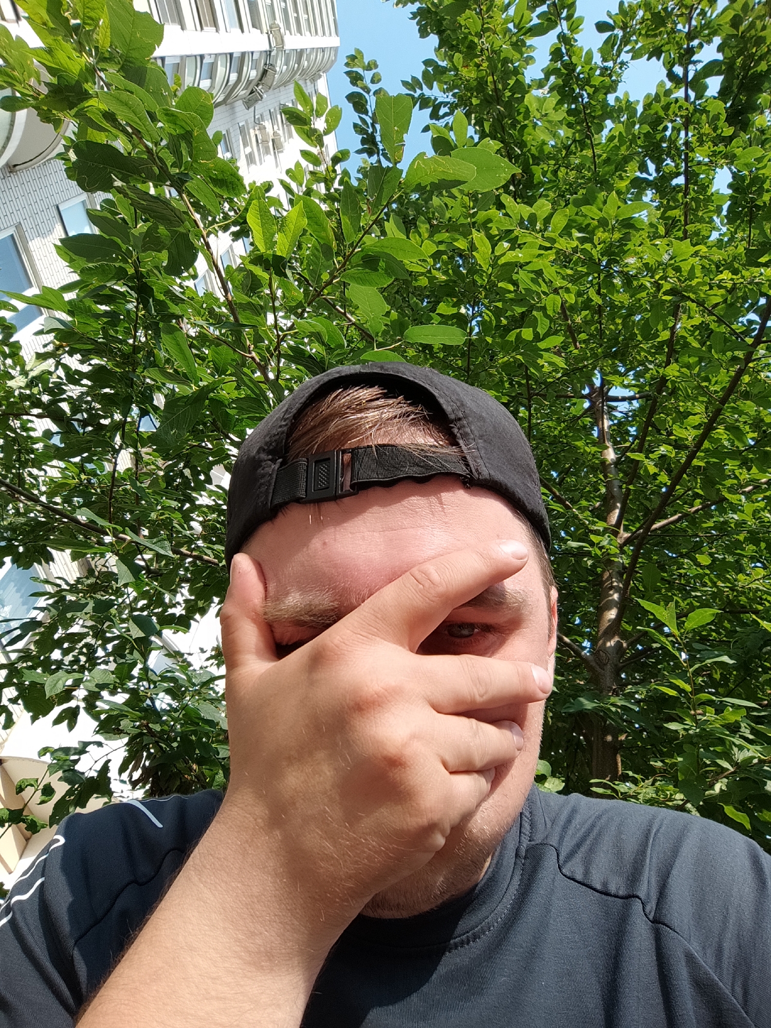 The image shows a person wearing a black baseball cap and a dark grey t-shirt, covering their face with their hand. The person appears to be outdoors with lush green foliage in the background, suggesting a park or a street lined with trees. Behind the trees, a modern white building with multiple floors and windows is visible, possibly an apartment complex. The sky is clear and blue. The lighting indicates it is daytime. The scene appears to be in an urban environment, with the building suggesting a city location. The presence of the building and foliage in this composition does not provide specific enough details to identify the exact location within the city of Vidnoye, Russia.
