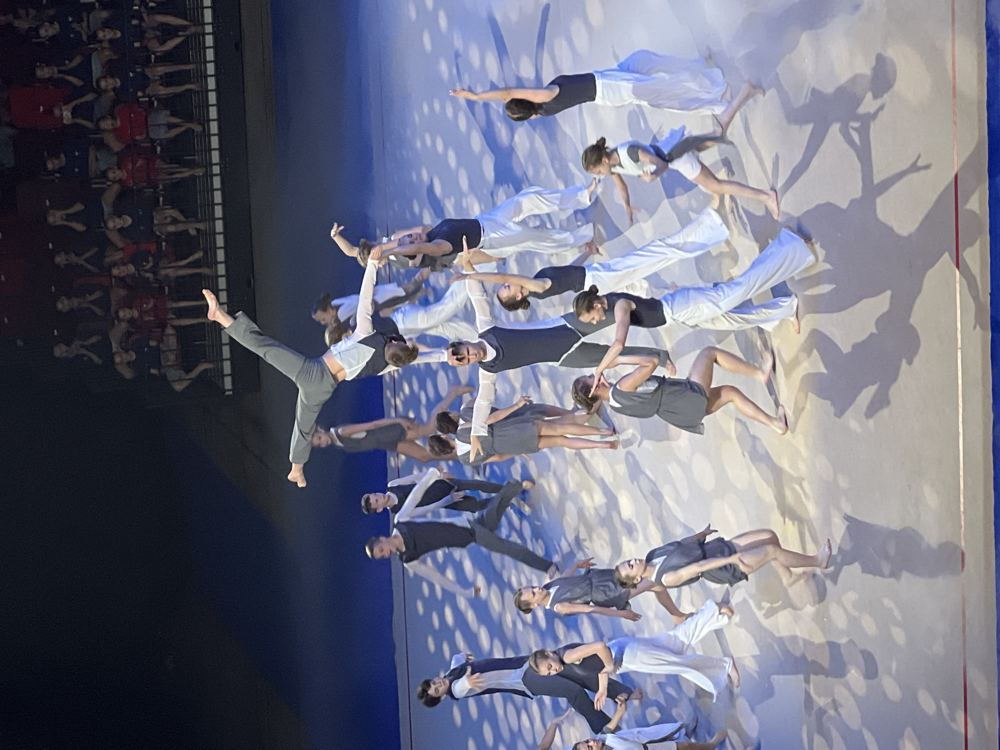 This is a high-angle, medium shot of a performance taking place on a stage with a blue backdrop and a white floor marked with circular patterns. A group of young performers, dressed in dark and white athletic attire, are engaged in a choreographed routine. The performers are in various poses, some are dancing, while others are in acrobatic positions, including a person being held upside down by another performer. In the background, spectators are seated in bleachers. The scene appears to be indoors, likely a theater or arena. The city of Moscow, Russia is not explicitly depicted or identifiable within the image.