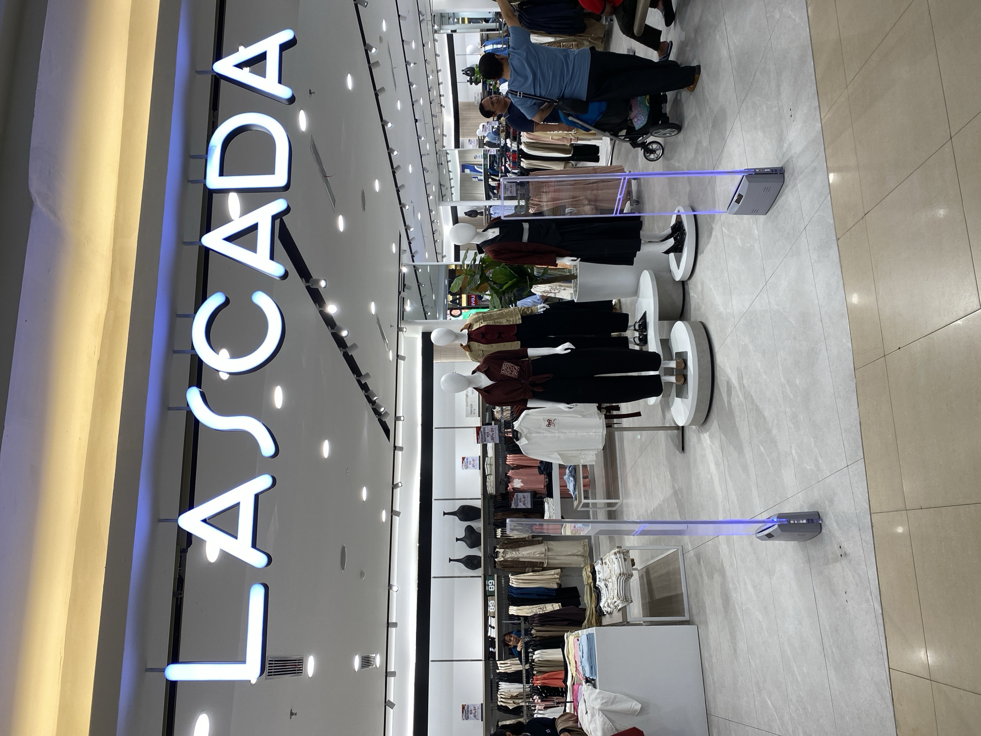 The image shows the exterior of a clothing store called LASCADA. The store has a prominent neon sign displaying the brand name in white letters with blue accents. In front of the store, there are several mannequins dressed in the latest fashion. One mannequin on the right wears a maroon top with a knot detail and black pants. Another mannequin in the center wears a maroon cardigan over a black dress. To the left, two mannequins are visible, one in a khaki jacket and black pants, and another partially visible in a maroon top and black pants. There are also displays of folded clothing and racks of clothes inside the store. A couple of security gates with blue lights are visible at the entrance. People are seen entering and exiting the store. In the background, more clothing racks and shelves are visible. The floor is tiled with a marble-like pattern. The overall impression is that of a modern and well-organized retail environment. The location is identified as Banjarmasin, Indonesia.