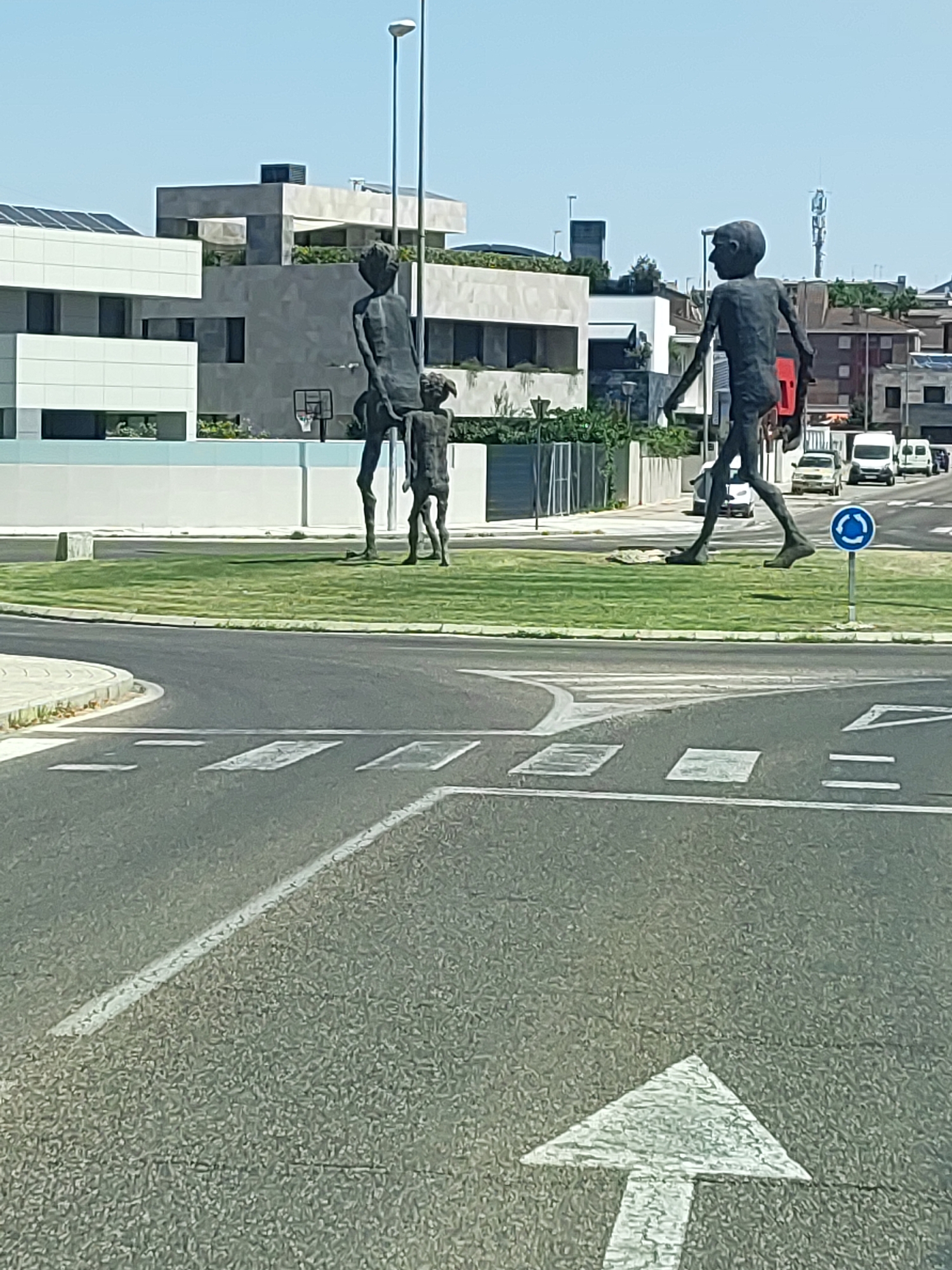 The image depicts a street scene in Palencia, Spain, featuring a roundabout with a prominent public art installation. The installation consists of three dark-colored, stylized human figures, appearing to be made of metal. Two larger figures, one walking to the right and another standing and facing left, flank a smaller figure positioned between them. Behind the sculptures, modern buildings with flat roofs and multiple levels are visible, some featuring solar panels. A basketball hoop with a backboard is also discernible in the background. The foreground shows a paved road with white lane markings, including a pedestrian crossing and an arrow indicating direction. Several vehicles, including vans and cars, are driving on the road in the background. A blue circular traffic sign with a white arrow indicates the direction of traffic in the roundabout. The sky is clear and blue, suggesting a sunny day.