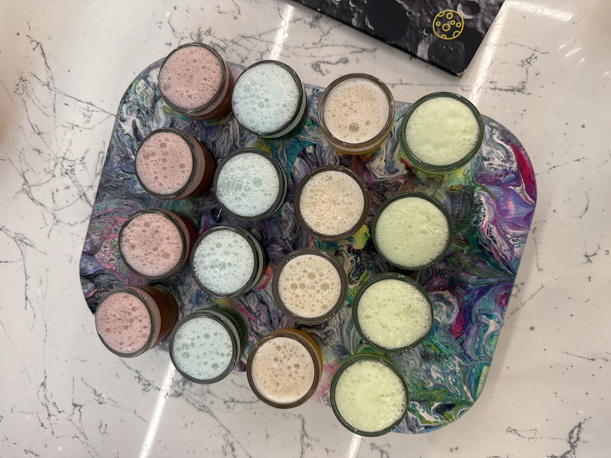 The image shows a top-down view of a colorful, swirled tray with 14 small glasses filled with brightly colored beverages. The tray rests on a white surface with a gray marble-like pattern. The glasses are arranged in four rows. The top row has three pink drinks. The second row has two light blue drinks. The third row has two light beige drinks and one light blue drink. The fourth row has four light green drinks and one light beige drink. The background shows a black book with a yellow circular emblem featuring craters. There are no people visible in the image. The scene is static and appears to be an indoor setting, possibly a bar or a cafe. The location in Saint Petersburg, Russia, is not discernible from the visual information provided in the image.