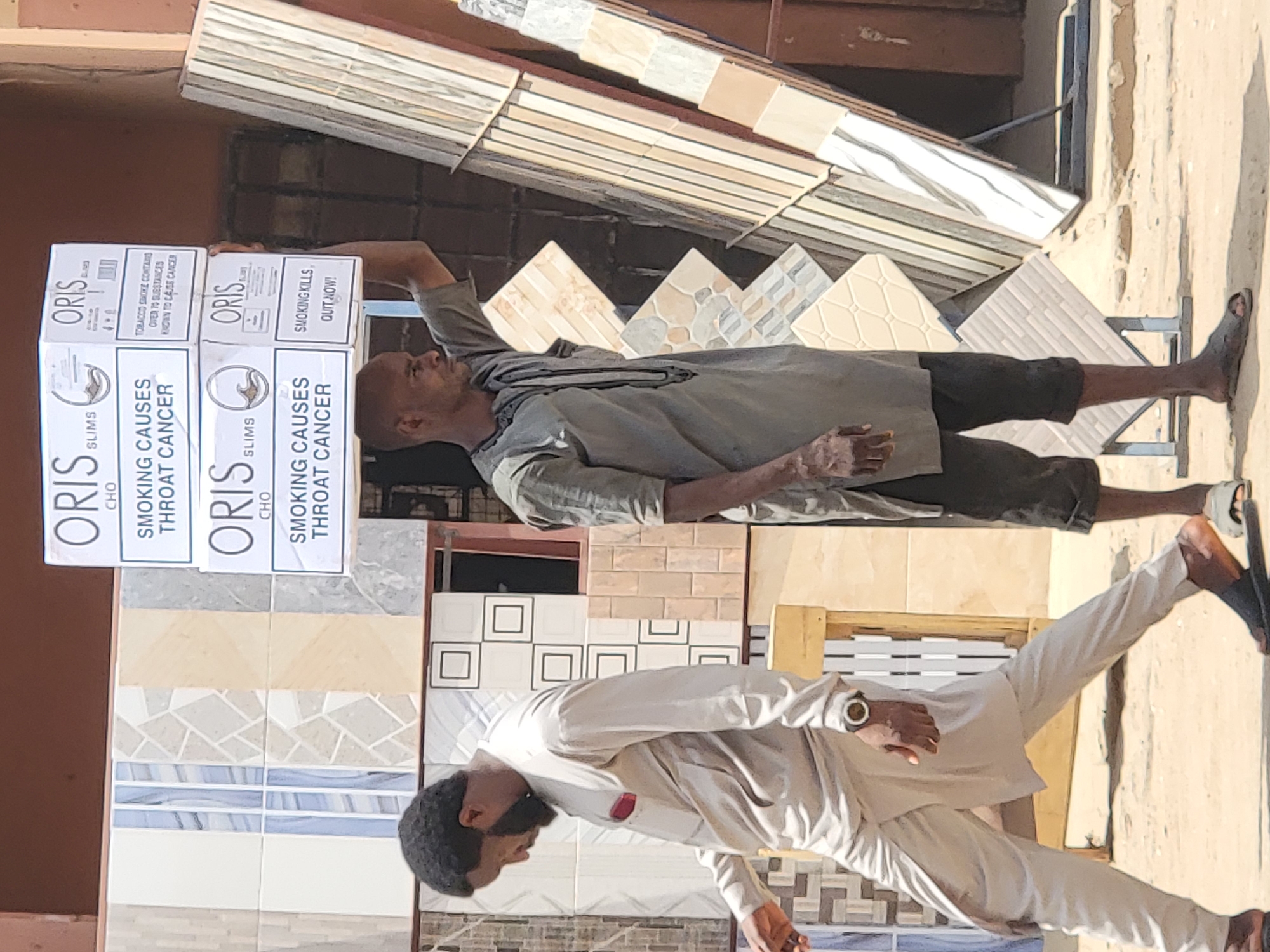 The image depicts a street scene in Maiburiji, Nigeria, featuring two men standing in front of a display of various tile samples. One man is holding a stack of cigarette cartons labeled "ORIS SLIMS" with health warnings in English ("SMOKING CAUSES THROAT CANCER," "SMOKING KILLS"). The environment includes a building or shop facade with a collection of different tile designs. The lighting suggests a sunny day. The men are interacting with the tile display.