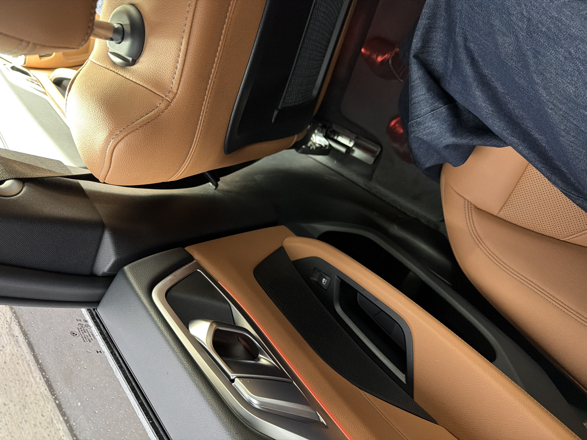 The image depicts the interior of a modern BMW vehicle, specifically focusing on the rear passenger compartment, observed from a forward-facing perspective. A person is seated in the rear, evidenced by their lower legs and feet.
**Subjects/Objects:**
* **Vehicle Interior:** The predominant colors are tan/light brown leather and black trim. The door panel on the left features tan leather, a chrome-finish door handle, a horizontal red ambient light strip, a black textured speaker grille, and a black power window switch.
* **Window:** A side window, positioned top-left, is wet with numerous raindrops, indicating precipitation. A faint BMW logo and text "43R-000492," "PPC PA-GF10," and "I 005080" are visible etched into the glass, likely safety markings.
* **Seats:** Two tan leather seats are visible. The rear seat, where the subject is positioned, features perforated leather on its base. The back of the front passenger seat includes a black mesh storage pocket.
* **Person:** The lower half of a person's body is visible, seated in the rear. They are wearing dark blue denim jeans. Two red objects, consistent with footwear, are on the floor beneath the front seat, dimly illuminated by a red light source on the floor.
* **Other Details:** Exposed metal brackets are visible on the floor beneath the front seat. The overall design suggests a contemporary vehicle with premium features.
**Actions/Interactions:**
* A person is occupying the rear passenger seat. No specific actions are observable beyond this occupancy.
**Environment:**
* The exterior environment is rainy, as indicated by the water droplets on the window. The vehicle appears to be stationary or moving slowly, observed from the interior.
**Location:**
* The scene is located in the city of Yerevan, Armenia.
