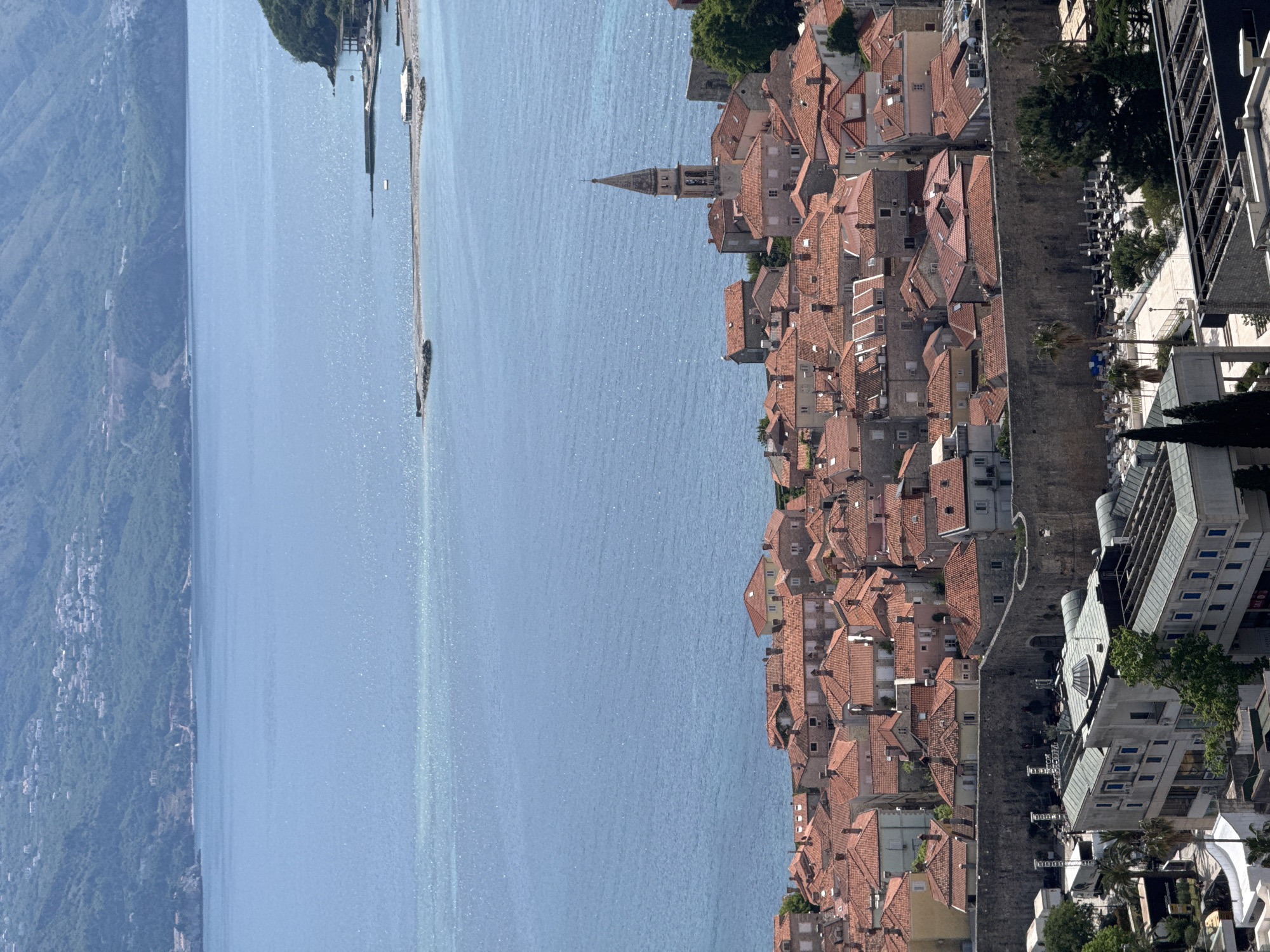 The image shows a view of Budva, Montenegro, overlooking the Adriatic Sea. Dense rows of buildings with terracotta roofs line the coast. A church steeple is visible among the buildings. Mountains are visible in the distance on the opposite side of the water. A pier or breakwater extends out into the sea. A modern multi-story building stands beside the old town, near the coast, complete with outdoor seating.