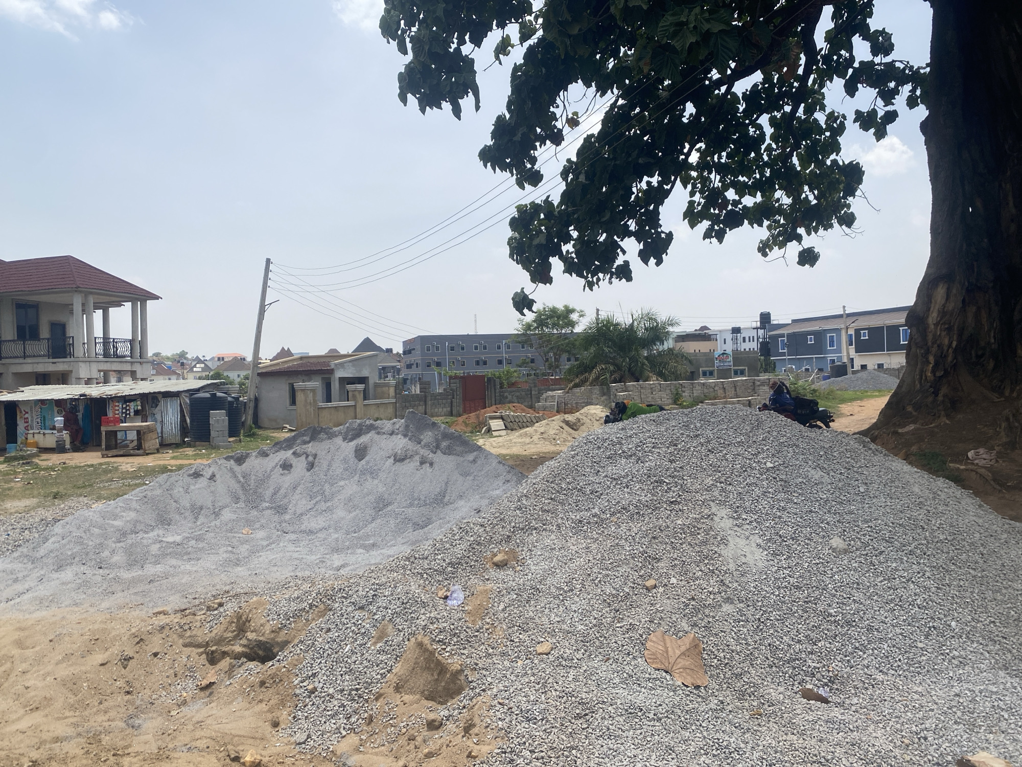 This outdoor, wide-angle image captures a semi-urban or suburban scene in Abuja, Nigeria, characterized by significant piles of building materials under a bright, clear sky. In the foreground, two large mounds of light grey gravel dominate the center and right, accompanied by a smaller pile of sand. A prominent, thick-trunked tree with dense green foliage frames the right side and top center of the image.
To the far left, a makeshift shop or kiosk with a corrugated iron roof displays items, and faint text, including "ABY" and "MILK", is visible on a cooler. Adjacent to it is a two-story residential building with a reddish-brown roof and a balcony featuring decorative railings. Black water tanks and concrete blocks are also visible near the shop.
A utility pole with power lines runs through the mid-ground, connecting to various buildings in the distance. A person is visible sitting on a motorcycle, partially obscured by the larger gravel pile, beneath the shade of the tree on the right. The background features a mix of residential structures, including a distinctive long, grey and blue building with square windows. The bright, slightly hazy daylight suggests it is daytime, likely morning or afternoon. The overall scene reflects a developing area with ongoing construction or material supply operations.