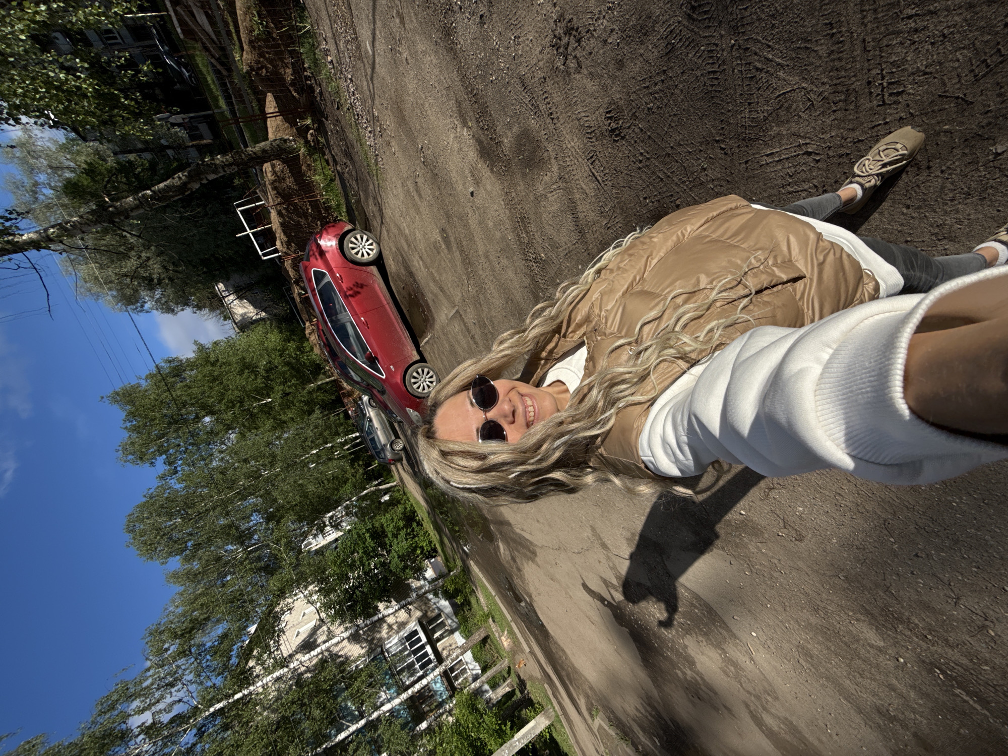 A selfie-style, tilted, upward-angle view in Rusanovo, Russia, shows a fair-skinned woman wearing round sunglasses, a tan puffer jacket, a white hoodie, and grey pants. She is smiling at the camera. Her blonde hair is wavy and long. She is standing on a dirt-covered road, casting a shadow from the right side of the frame. A red car is parked to the left. Trees and buildings are visible in the background under a blue sky with scattered clouds. The scene is well-lit.