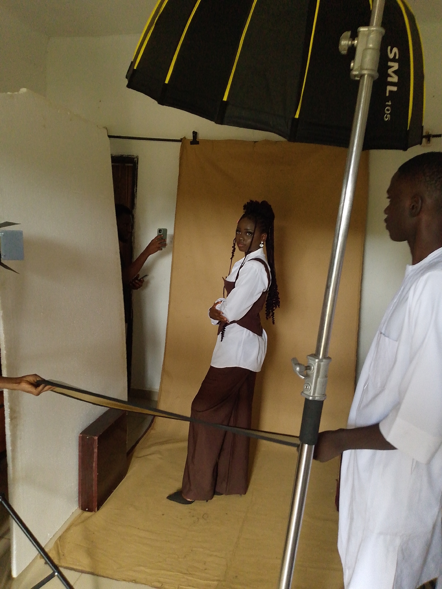 In a studio in Bukuru, Nigeria, a young Black woman with braided hair is posing for a photoshoot. She is wearing a white blouse with a dark brown corset over it and brown wide-leg pants. She is standing in front of a tan backdrop, with a large umbrella lighting rig positioned above and to the right. To the right of the woman, a Black man in a white traditional outfit is standing, looking at her. In the background, another person is holding up a smartphone, seemingly taking a photo or recording a video of the photoshoot. The studio environment appears to be sparsely decorated with a few props visible, including a wooden shelf unit.