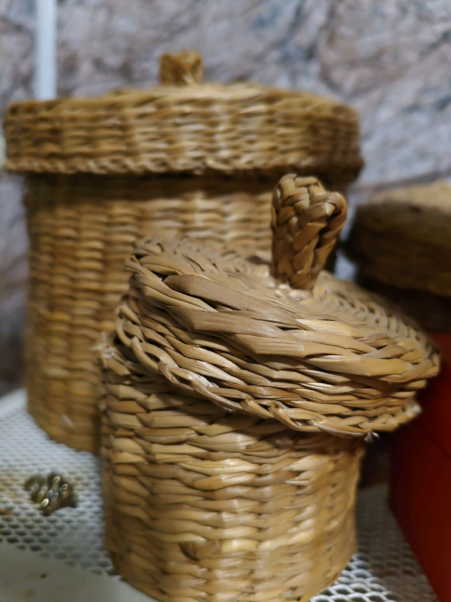 In Rostov-on-Don, Russia, two woven baskets are displayed on a white surface in front of a textured wall. The basket in the foreground is smaller and has its lid partially open, revealing a braided handle. Behind it, a larger, lidded basket stands to the left. To the right of the foreground basket, a portion of a red object is visible. A few small, shiny objects, possibly jewelry or beads, are scattered on the white surface to the left of the foreground basket. The lighting is dim, casting shadows and highlighting the woven texture of the baskets.
