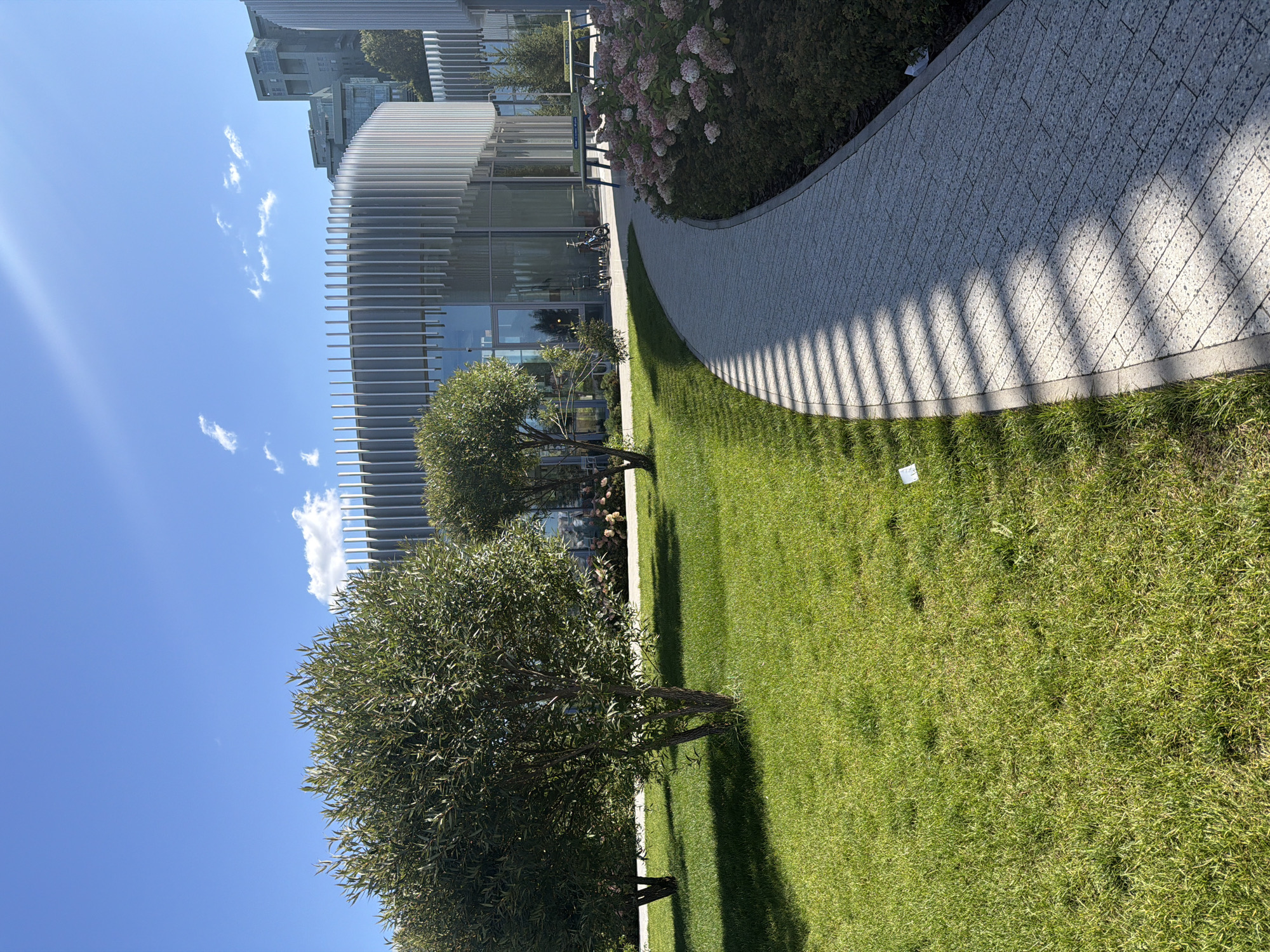 The image shows a modern architectural complex with a curved facade made of vertical metal slats and large glass windows. In the foreground, a paved walkway curves to the right, bordered by a lush green lawn and bushes with pink flowers. Several trees with dense foliage are scattered across the lawn. In the background, other buildings of similar modern design are visible, along with taller structures in the distance. The sky is clear blue with a few wispy clouds, and the sun is casting strong shadows. The scene appears to be a well-maintained public space or park within an urban setting. The provided information indicates this scene is located in Kazan, Russia.