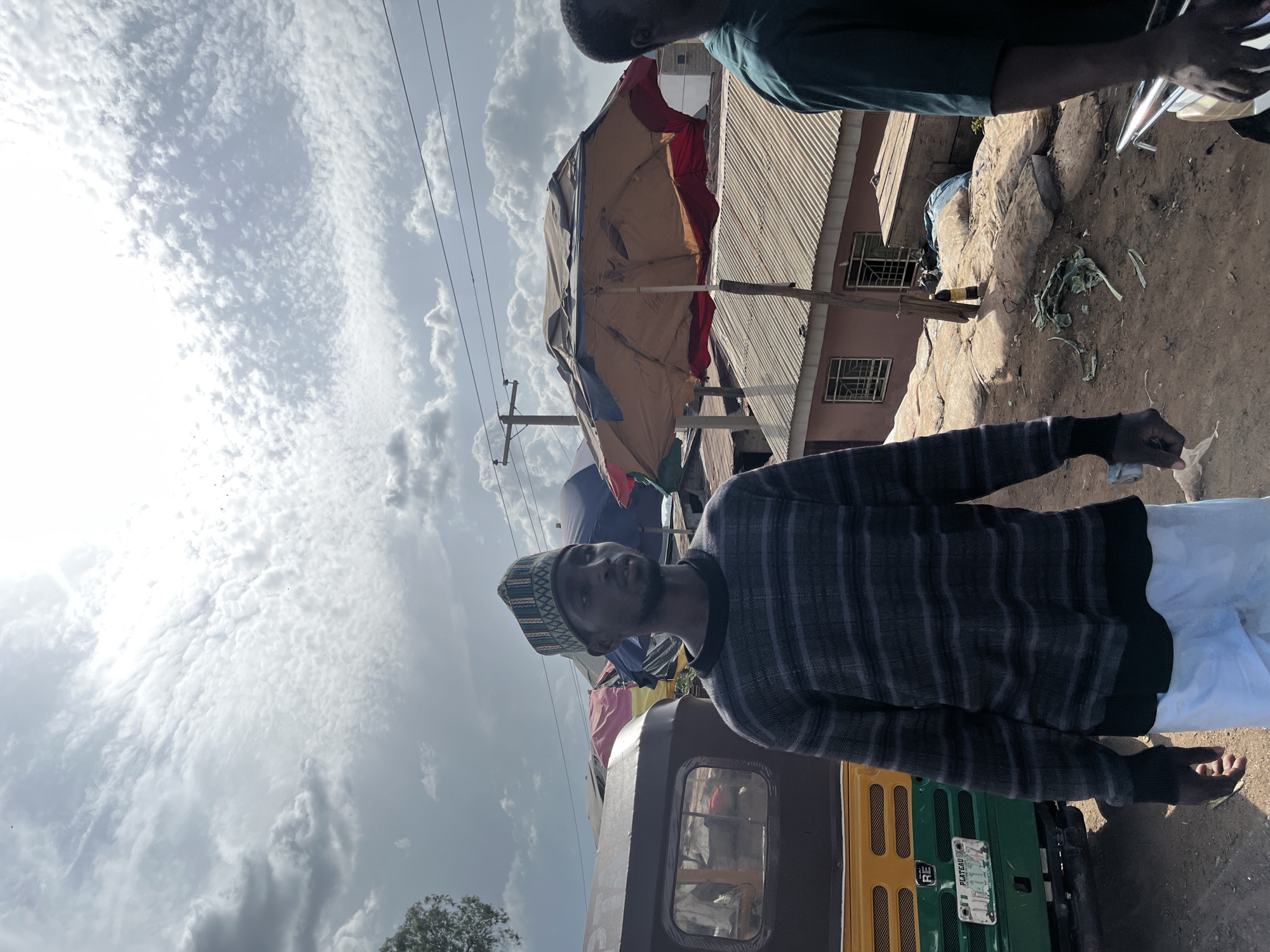The image depicts an outdoor scene in Jos, Nigeria, under a cloudy sky. The primary subject is a man wearing a striped sweater and a patterned cap, standing next to an auto-rickshaw. The auto-rickshaw is partially visible, showing its yellow and green colors, with a license plate that reads "PLATEAU RE".
In the background, there are buildings with corrugated metal roofs, suggesting a commercial or residential area. A large, colorful umbrella, likely for shade, is set up near the buildings. The ground is dirt and appears uneven. There are power lines visible in the sky, and some debris, including what looks like dry vegetation, on the ground.
The lighting suggests it is daytime, possibly late morning or afternoon, given the angle of the sun and the presence of shadows. The weather appears overcast with scattered clouds. The man's expression is neutral as he looks upwards. The scene appears to be a marketplace or a street scene with some vendors or shopfronts. There is no discernible activity beyond people standing around.