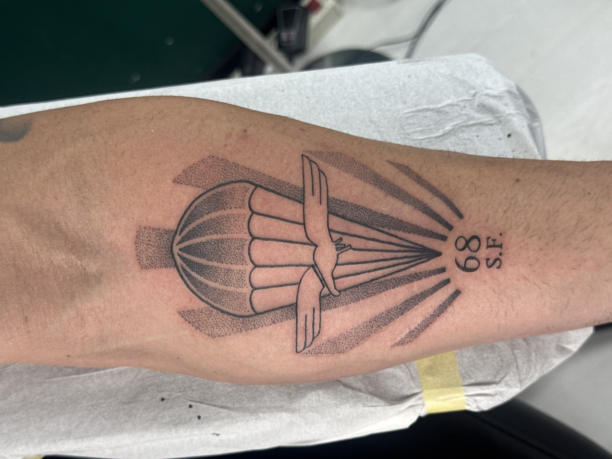 A tattoo on a light-skinned arm is visible. The tattoo features a parachute with lines emanating from it. Superimposed on the parachute are two hands. To the right of the parachute is the text "68 S.F." The arm is resting on white paper that has been taped down with yellow tape. In the background is a blurry image of what appears to be medical equipment. The photo appears to be taken indoors.