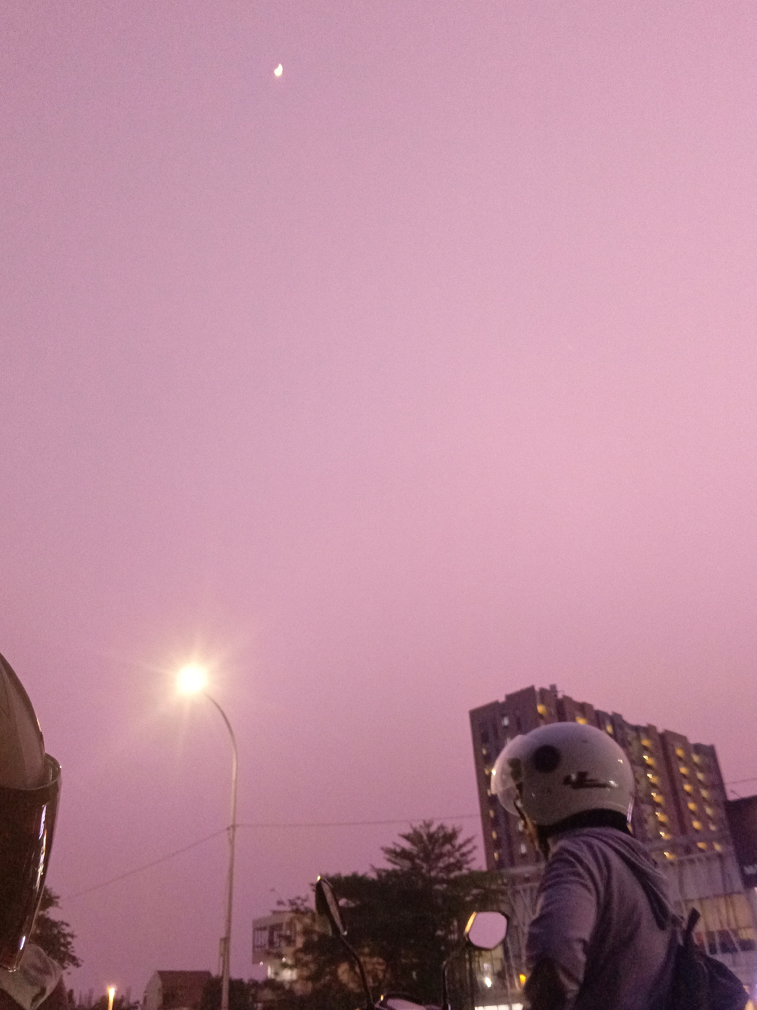 A low-angle, wide shot captures a dusky sky with a faint crescent moon visible in the upper left quadrant, set against a soft pinkish-purple atmosphere. In the foreground, the upper portion of a person wearing a light-colored helmet and a grey hooded jacket is visible on the right, looking towards the right. Below and to the left, another helmet, with a dark sheen and hints of brown, is partially visible. A street light with a bright bulb illuminates the scene from the mid-left, casting a glow. In the background, a multi-story building with illuminated windows stands prominently on the right, suggesting an urban setting. Trees and other buildings are faintly discernible amidst the soft light. The scene appears to be in the city of East Jakarta, Indonesia.