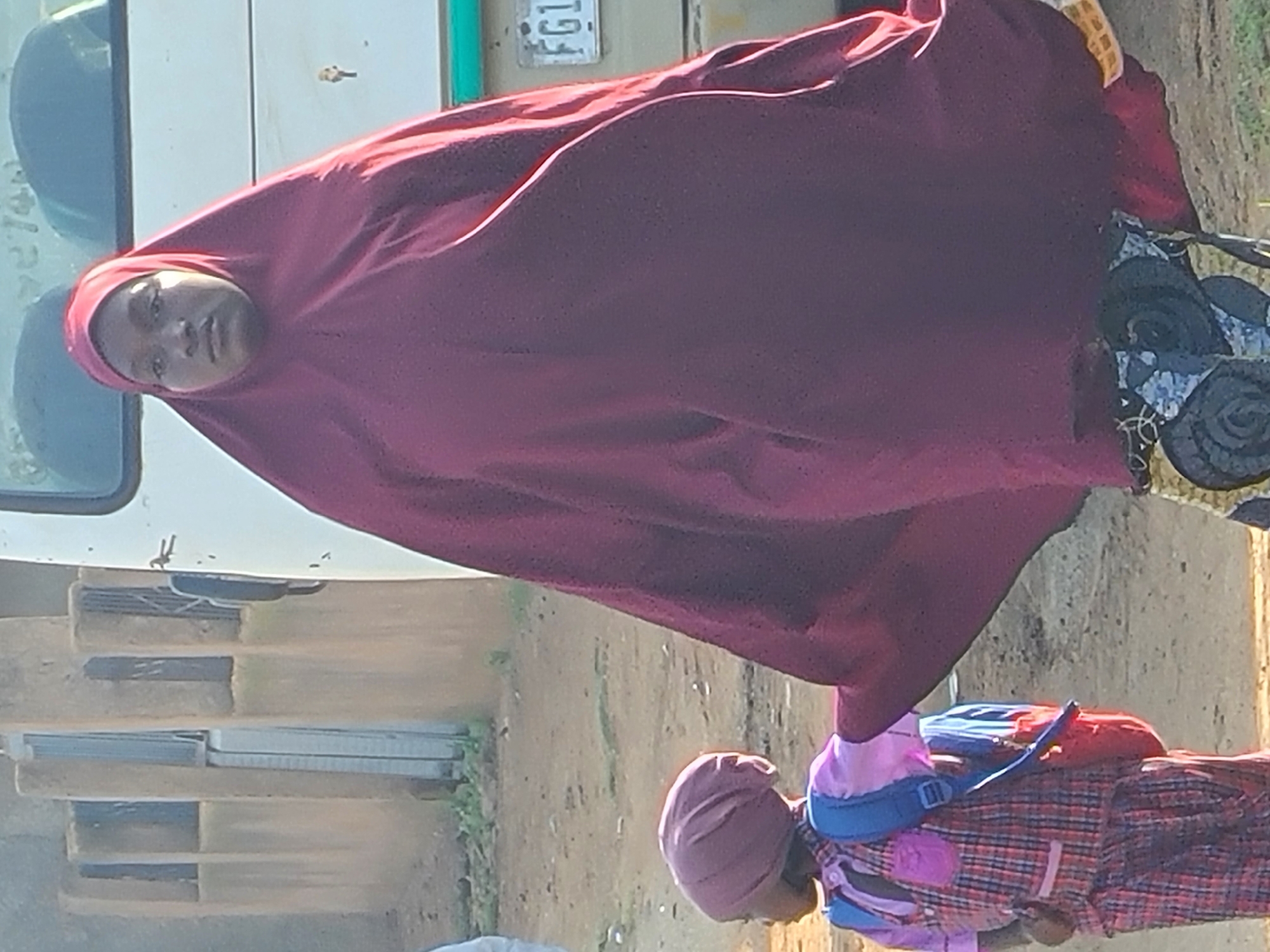 A young girl wearing a pink and red plaid dress and a purple headscarf stands to the left of a taller person wearing a burgundy abaya and headscarf. The girl has a blue backpack with a red pouch attached. They are on a dirt path in Jos, Nigeria. Behind them is a building with vertical slats and a white vehicle with a license plate that reads "FG1".