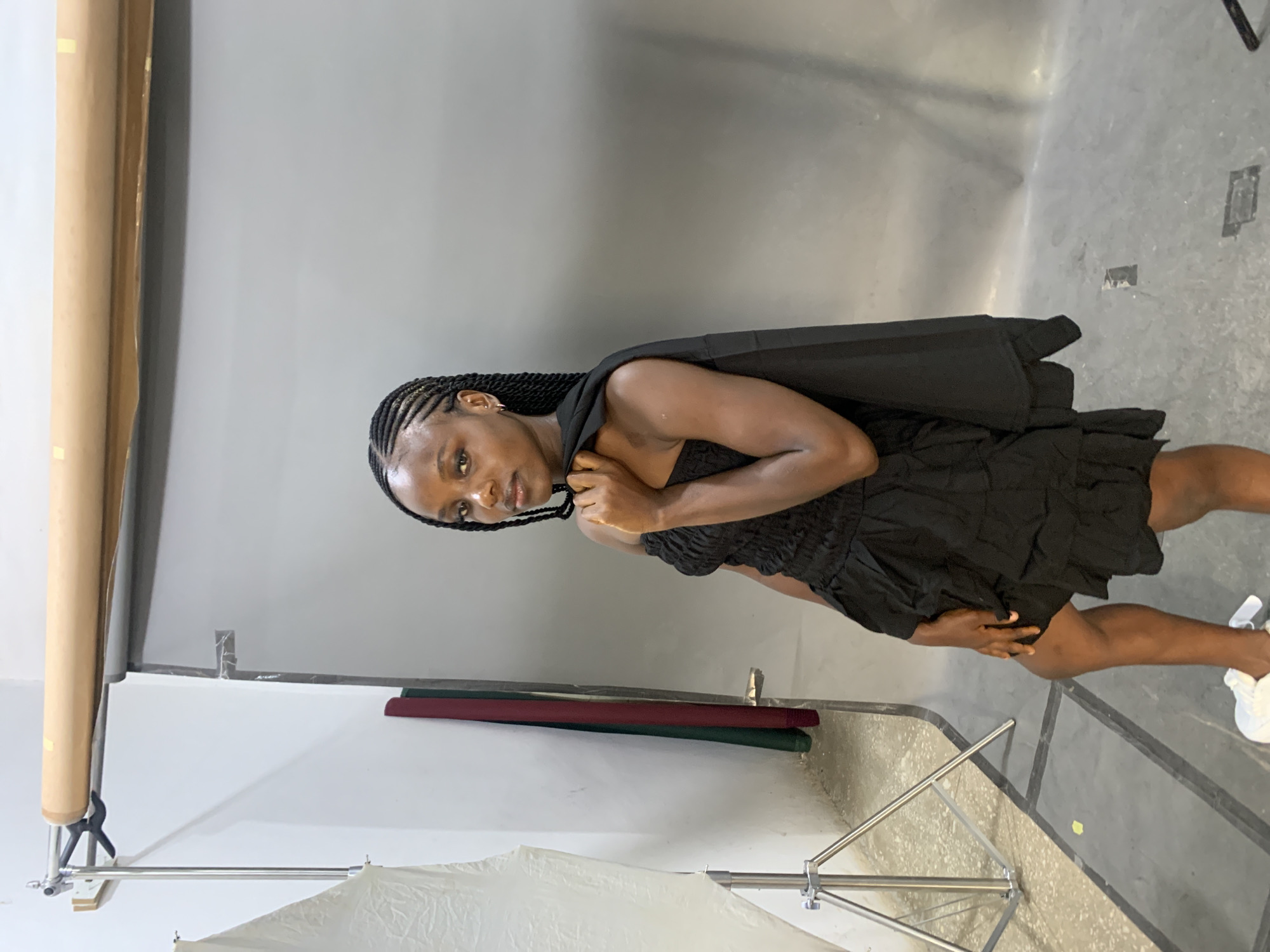 A young Black woman with braided hair stands in a photography studio. She is wearing a black strapless dress and a black jacket draped over her shoulders. Her right leg is bent and extended forward. She is holding the lapel of her jacket with her left hand. The background is a plain gray backdrop. To the left of the woman, there is a white reflector on a stand. There are also two rolled-up backdrops, one red and one green, leaning against the wall behind the reflector. The floor is concrete. The scene appears to be indoors. There is no specific information to suggest the location is in Gura Top, Nigeria.