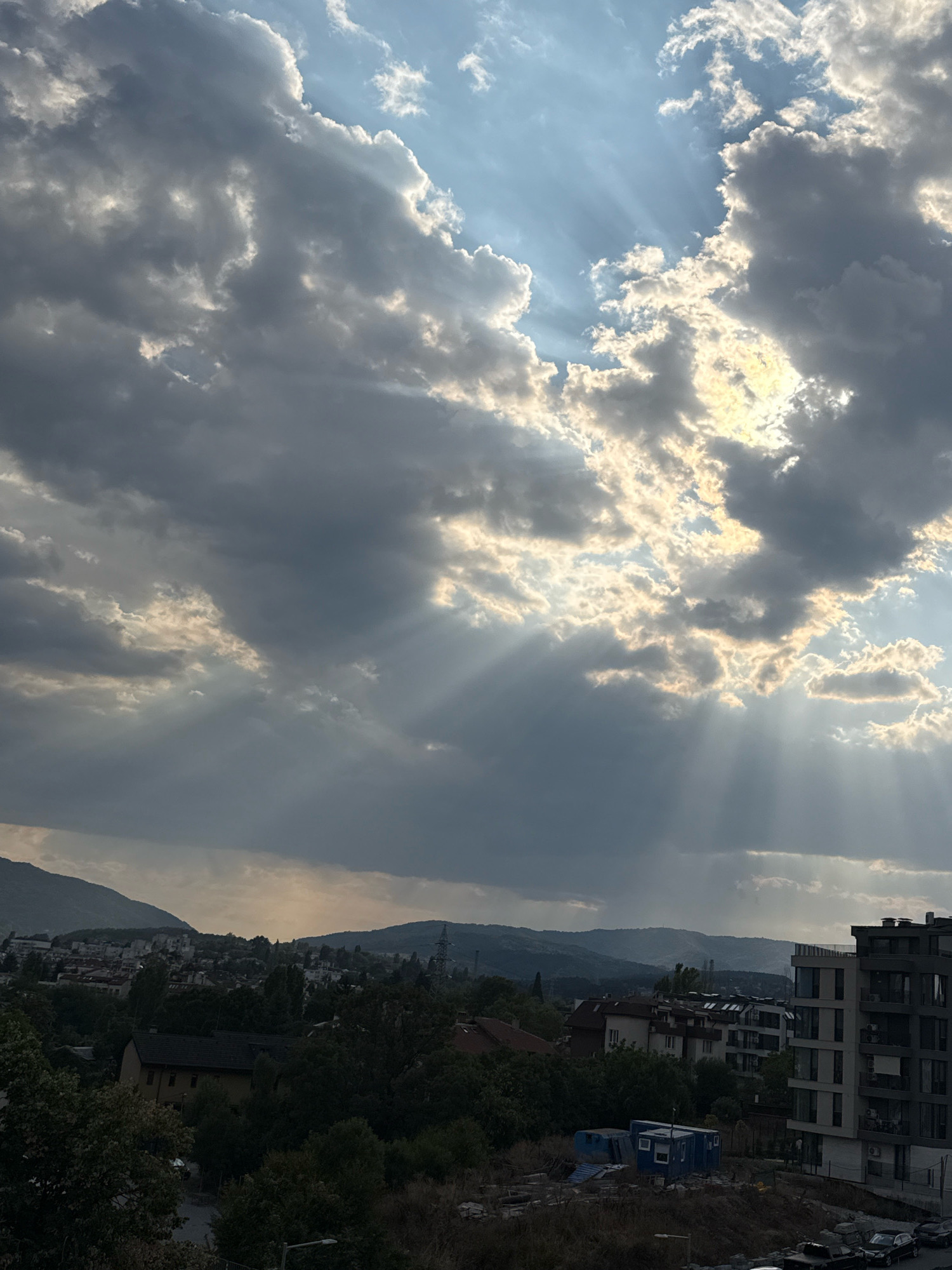 The image depicts a panoramic view of a city, likely Sofia, Bulgaria, during a cloudy day with dramatic sunbeams breaking through the overcast sky. In the foreground, there are hints of urban development with buildings of varying architectural styles, including a modern, multi-story apartment building on the right side of the frame, and what appear to be modular structures or temporary housing units further left. Trees and some shrubbery are visible, suggesting a mix of urban and natural elements. In the middle ground, residential buildings and other structures are nestled amongst trees, with a characteristic European feel. The background is dominated by a range of hills or mountains, shrouded in a light haze, characteristic of urban environments. The sky is a prominent feature, with large, dark clouds interspersed with patches of bright blue sky. Powerful rays of sunlight are piercing through the clouds, creating a striking visual effect known as crepuscular rays. The overall impression is one of a dynamic sky over a developing urban landscape.