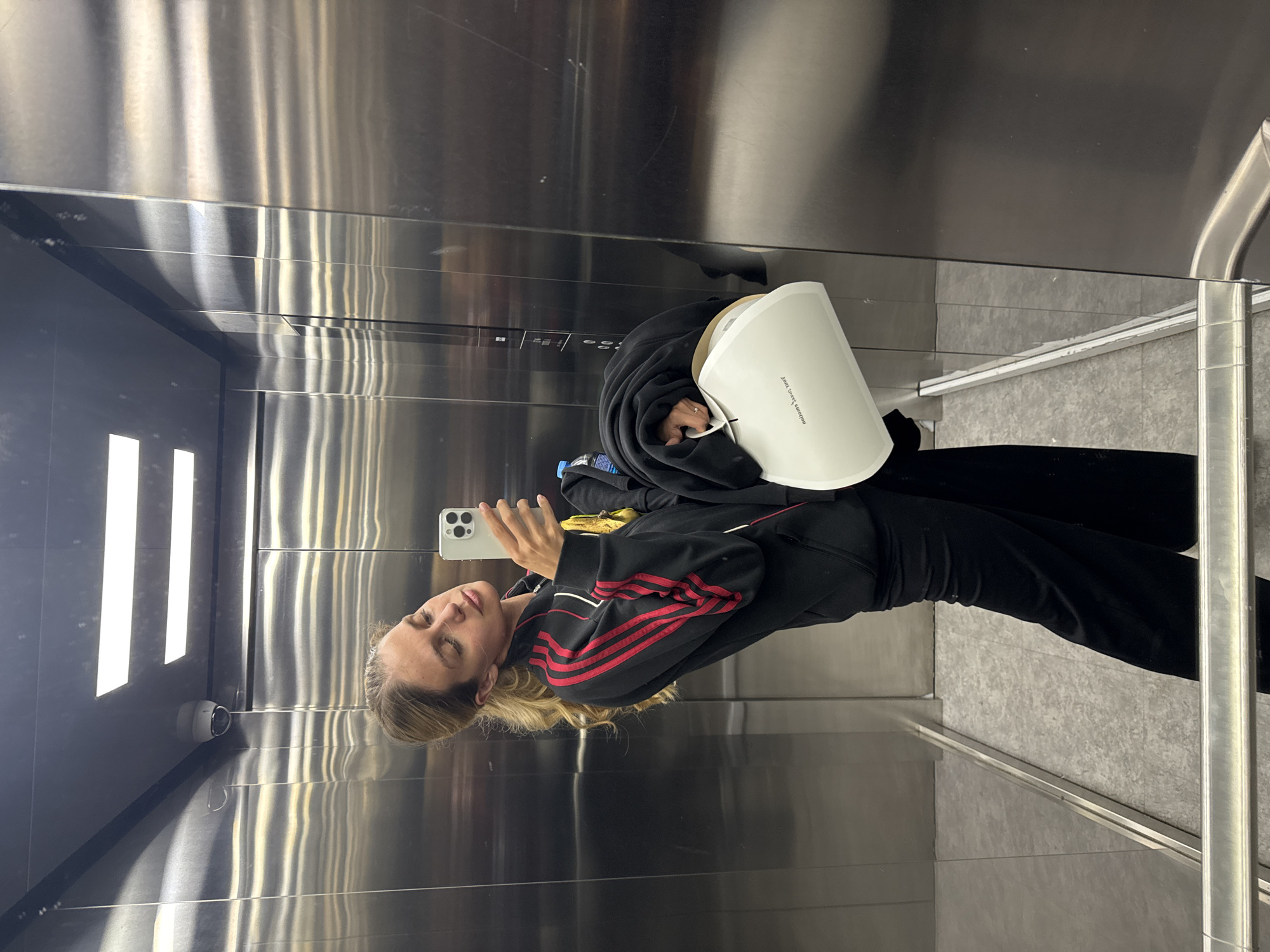 The image depicts a woman taking a mirror selfie inside an elevator. The elevator interior features highly reflective, brushed stainless steel walls that show vertical streaks of light and various reflections. The floor is a light gray, textured material, possibly tile or linoleum.
The woman, with blonde hair, is positioned in the reflected view, holding a light-colored smartphone with her right hand to capture the selfie. Her eyes appear to be closed or partially closed. She is dressed in a black long-sleeved top or hoodie with prominent red stripes running down the sleeves, along with black pants. Under her left arm, she carries a large, white, curved object, which looks like a folder or a unique bag. On this object, the reflected and inverted text "moment in time" is discernible. A blue-capped water bottle is tucked into her clothing on her left side.
Within the elevator's reflection, a white security camera is mounted on the upper left wall, and multiple vertical, rectangular light fixtures illuminate the space. A panel of elevator control buttons is visible in the reflection to the left of the woman. A metallic handrail extends along the right side of the elevator's interior. The lighting is bright and even throughout the scene.